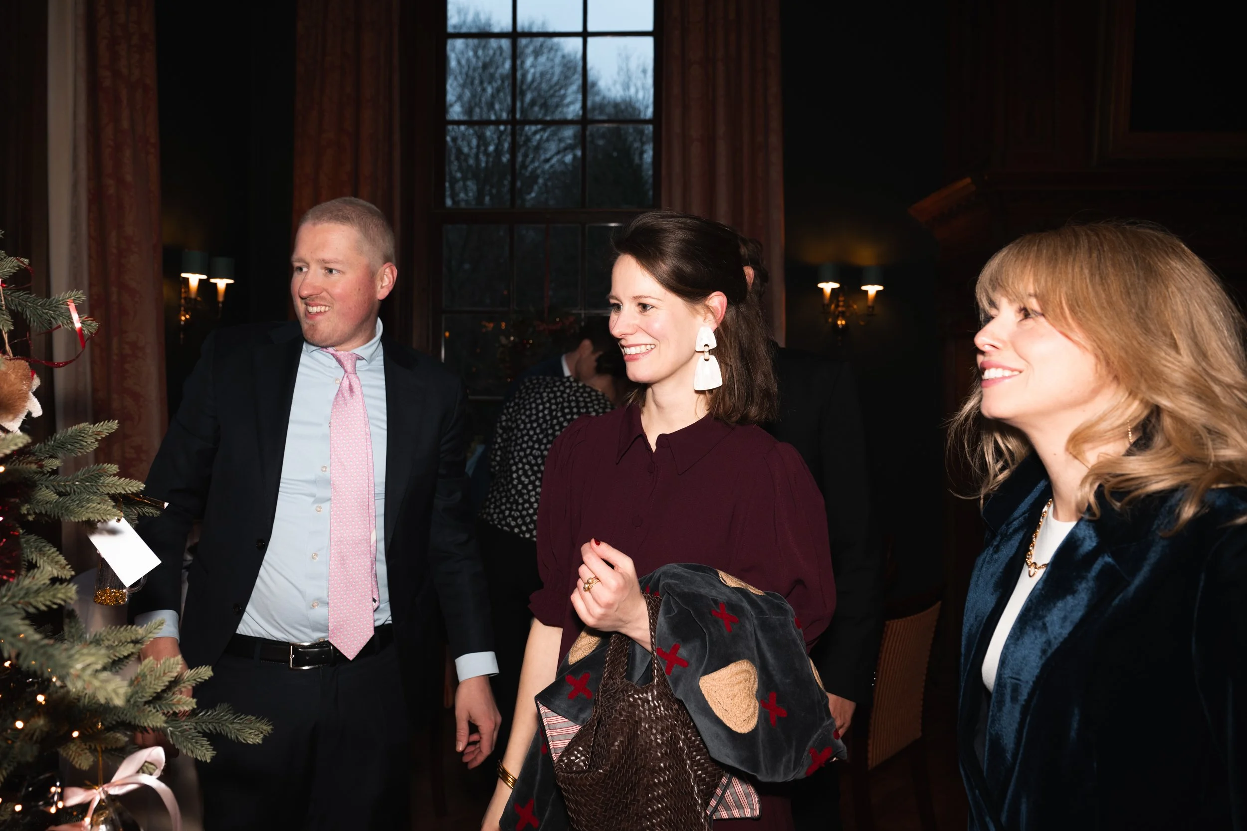 Three people, two women and one man, are standing near a decorated Christmas tree indoors, smiling and engaging in conversation.