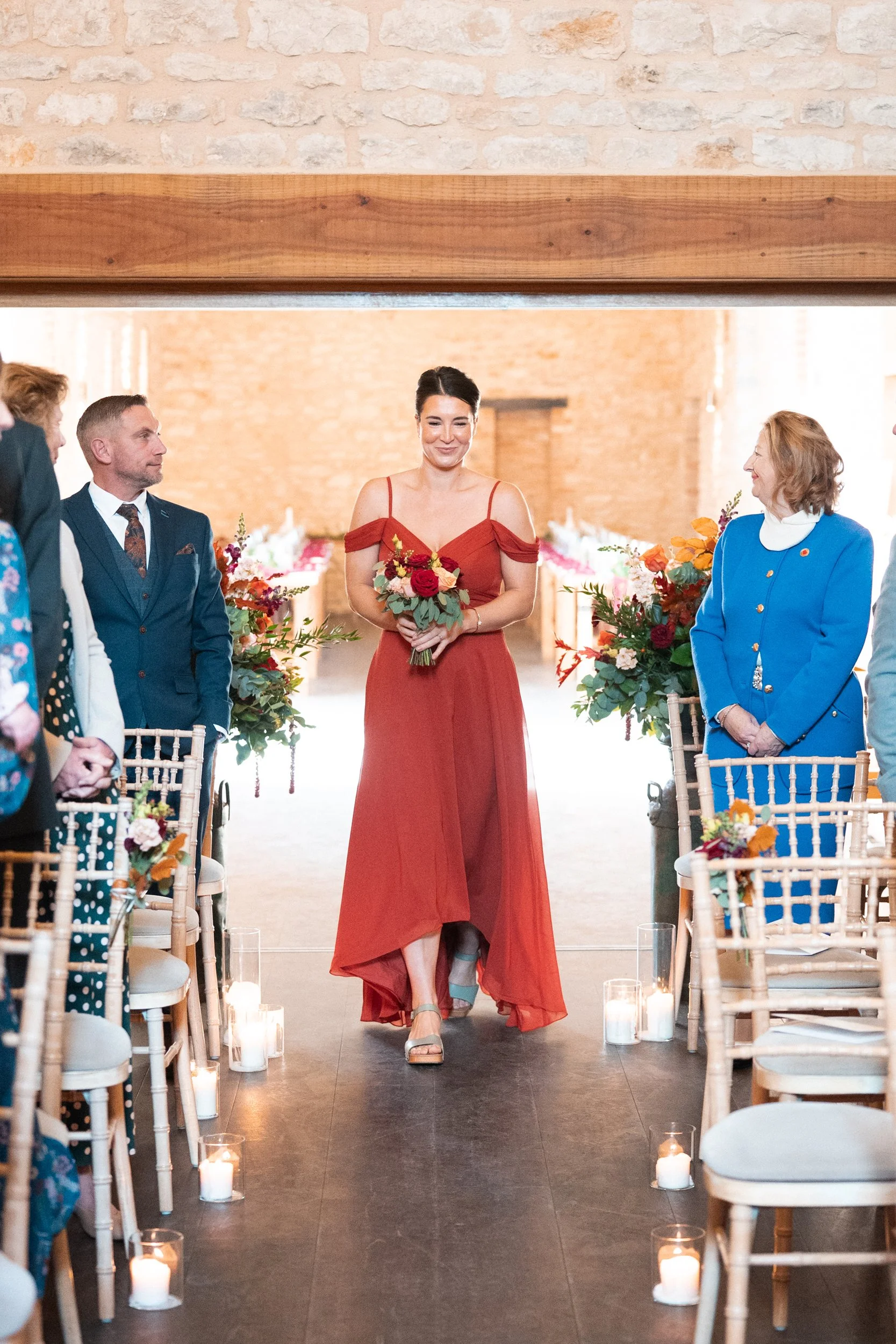 A woman in a red dress is walking down the aisle holding a bouquet, flanked by wedding guests on either side, with candles and floral arrangements along the aisle in a decorated venue with brick walls.