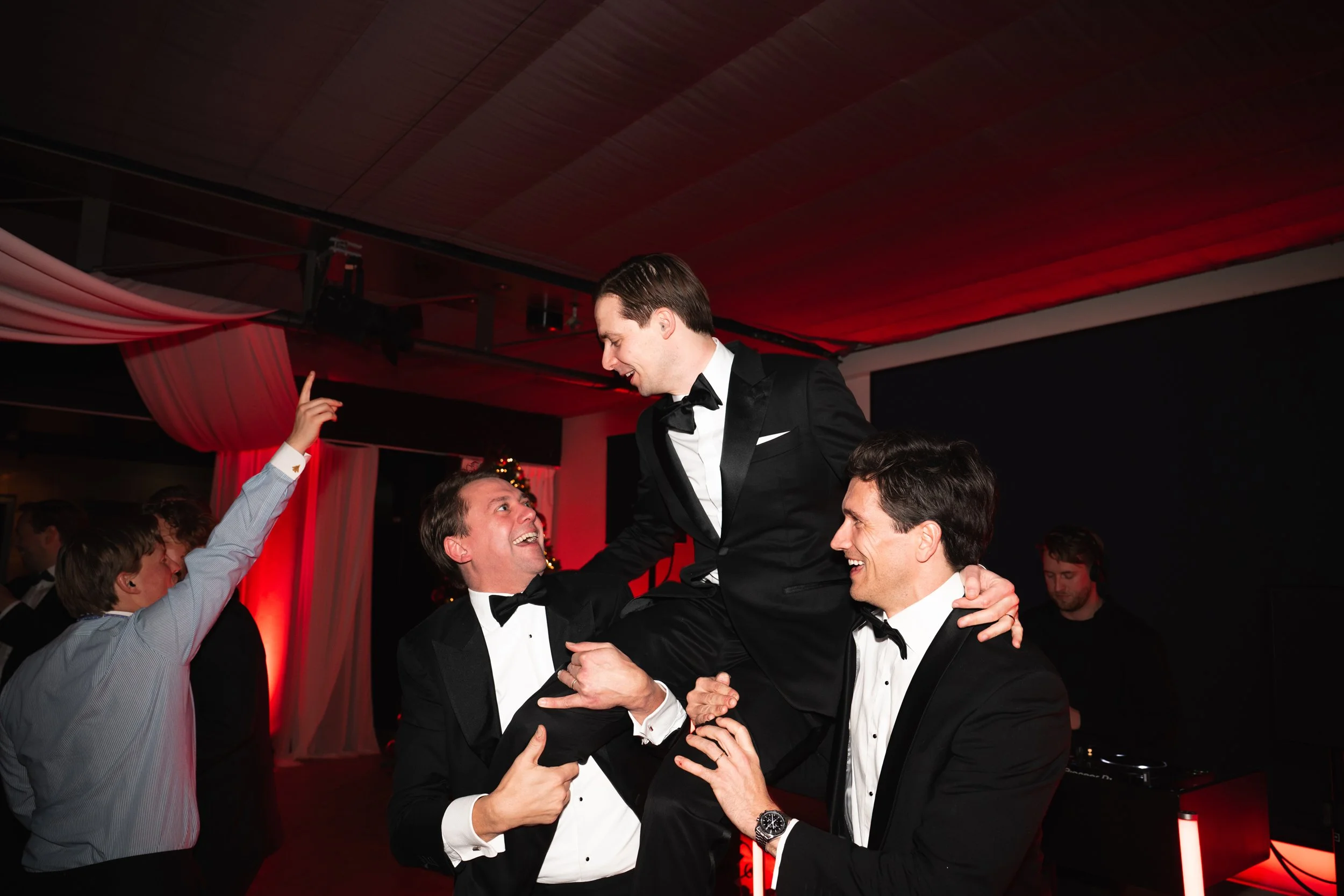 Men in tuxedos celebrating at a formal event, with one man lifted in the air by two others, as others dance in the background.