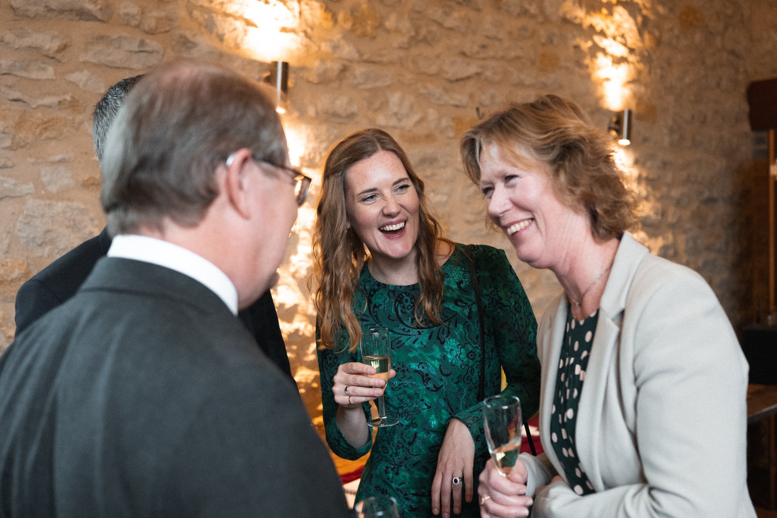 Group of four people, three women and one man, engaged in conversation at a social gathering, holding champagne glasses, with a stone wall background and warm lighting.