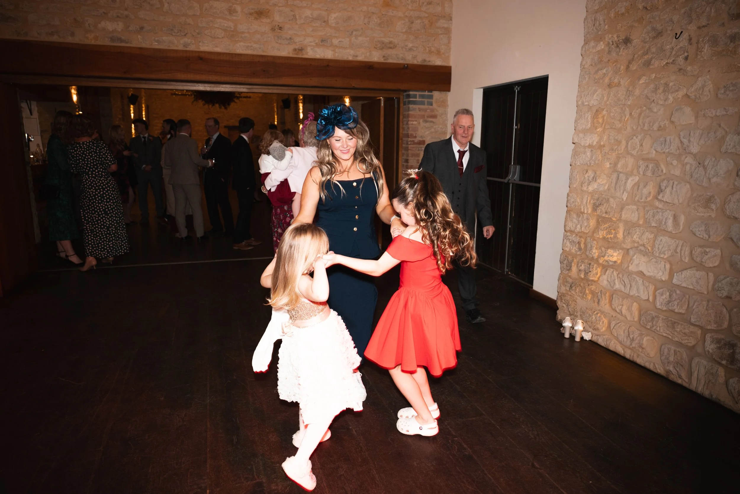 Two young girls dancing with a woman at a wedding reception in a rustic venue, with other guests mingling in the background.