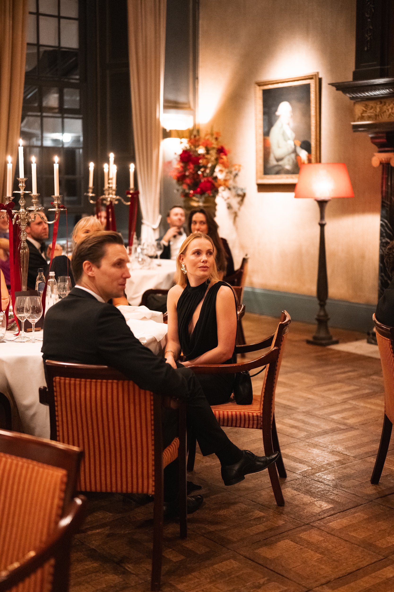 People seated at a formal dinner in a decorated, elegant restaurant with candelabra, framed portraits, and warm lighting.
