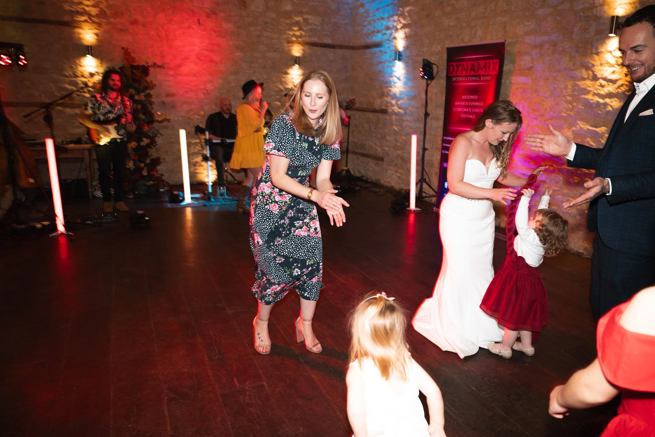 People dancing at a wedding reception with a band playing in the background, including a bride in a white dress, a man in a suit, and children in red dresses.