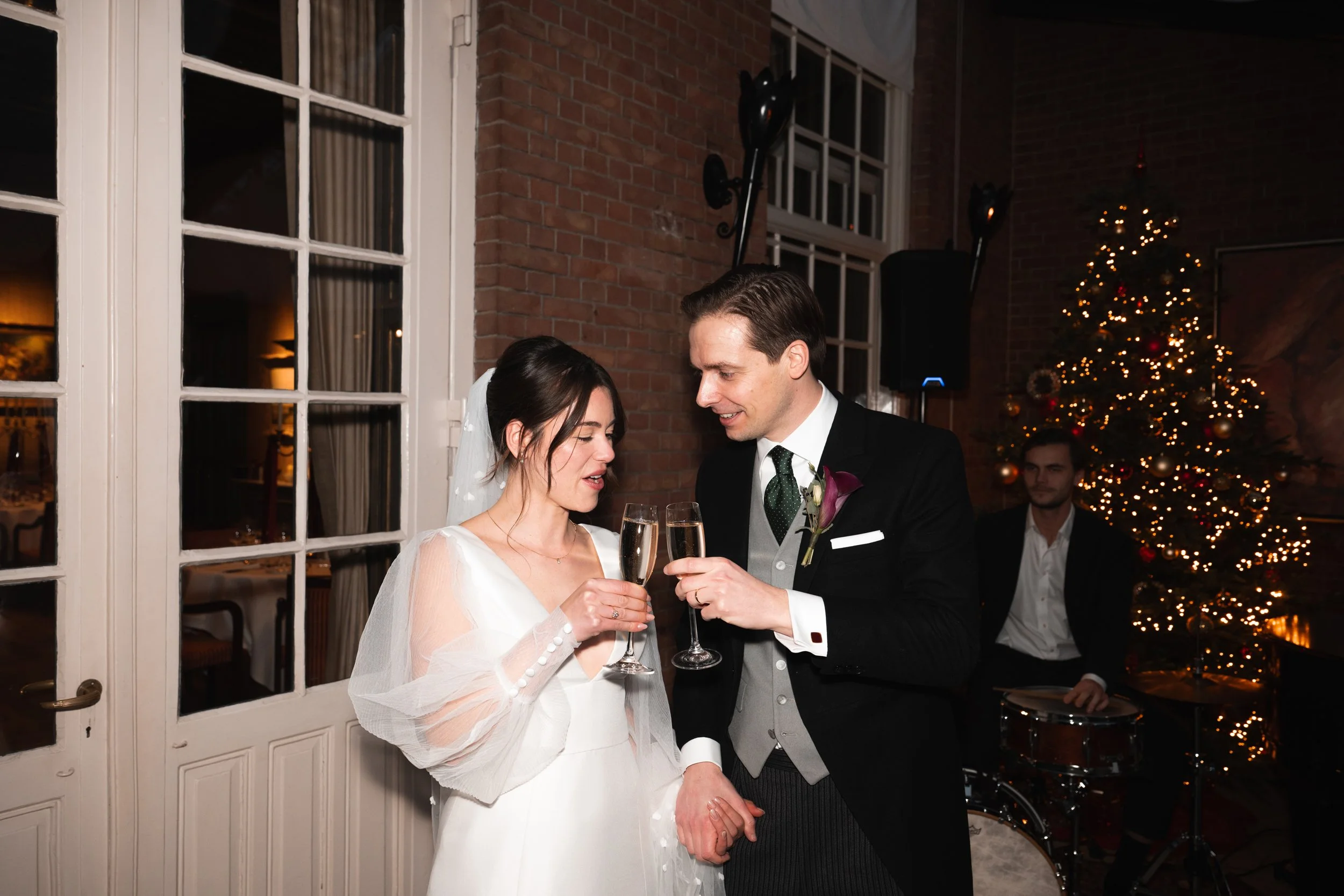 A bride and groom toasting with champagne glasses at their wedding reception, holding hands, with a Christmas tree and drummer in the background.