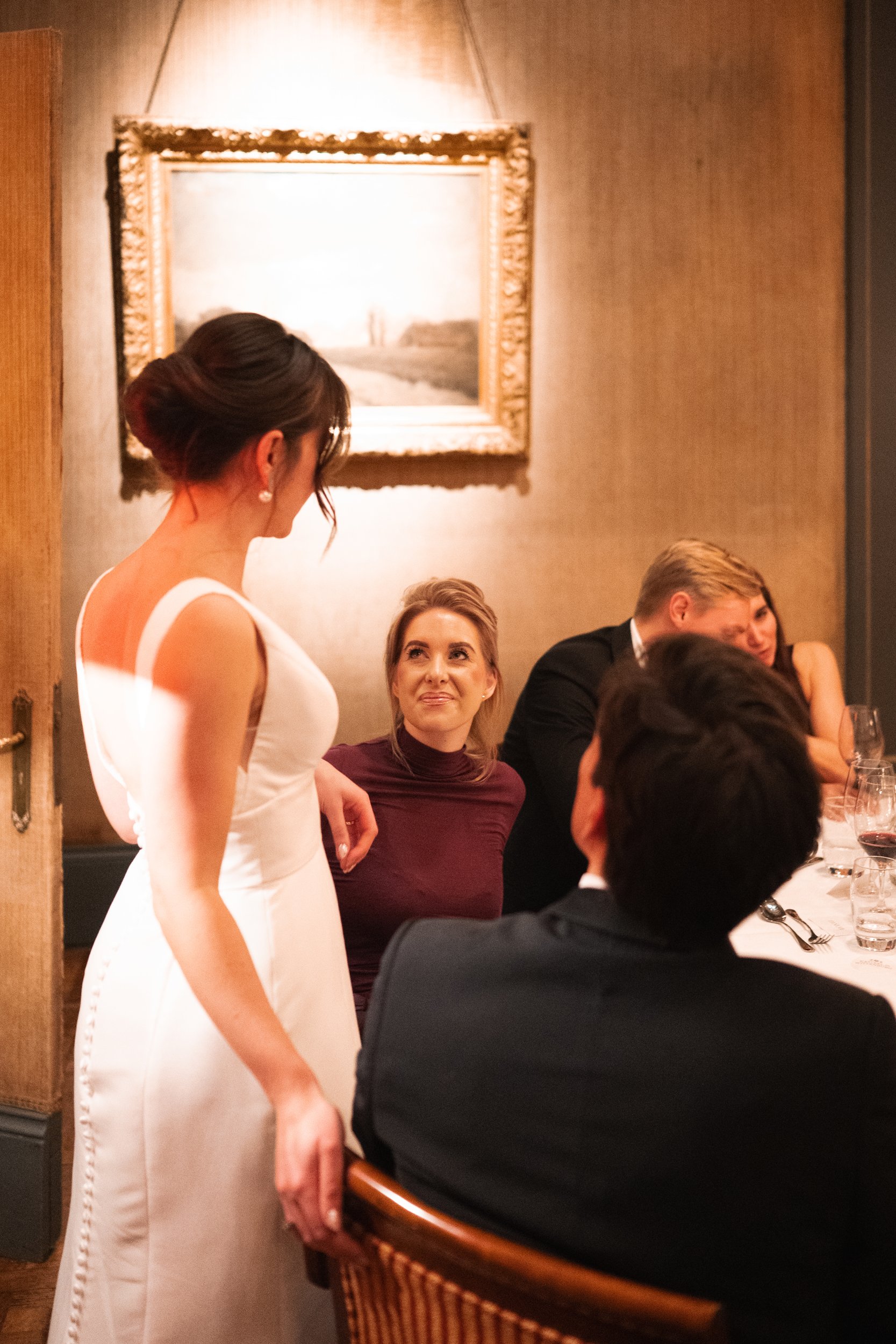 A woman in a white dress stands and talks to a group of seated people at a dinner table in an elegant restaurant.