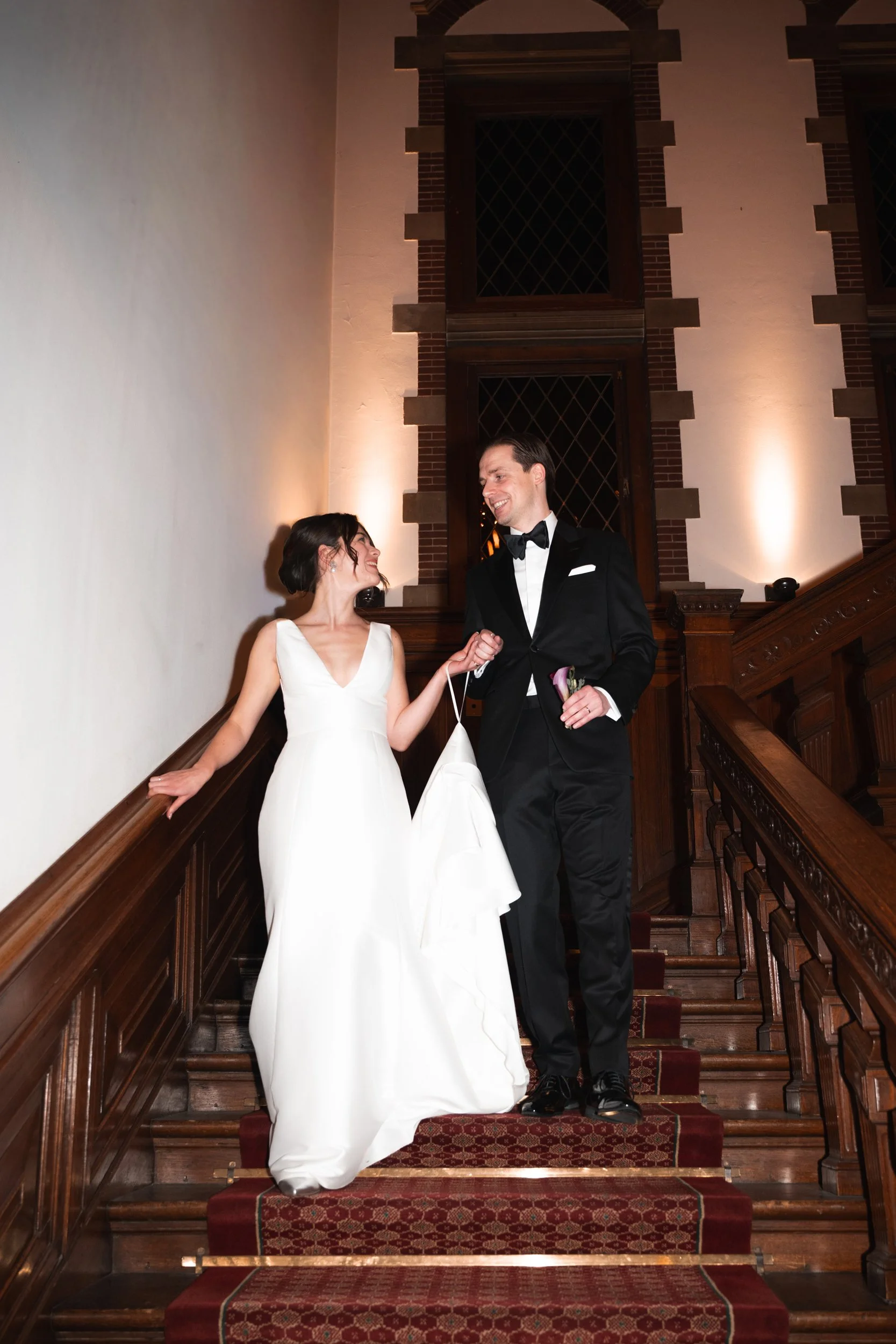Wedding couple on staircase, woman in white wedding dress, man in tuxedo, smiling at each other.