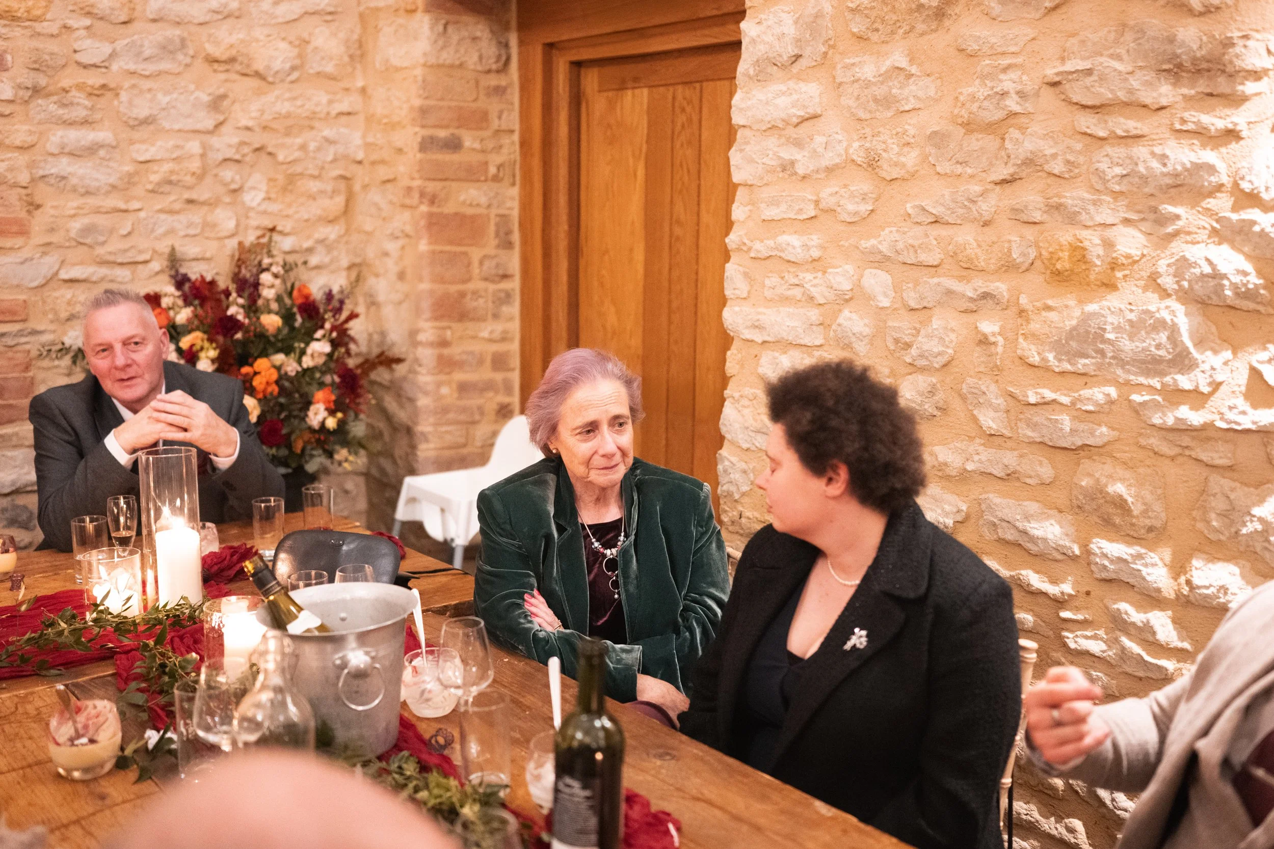 People sitting around a decorated dinner table inside a rustic stone-walled room, having a conversation.