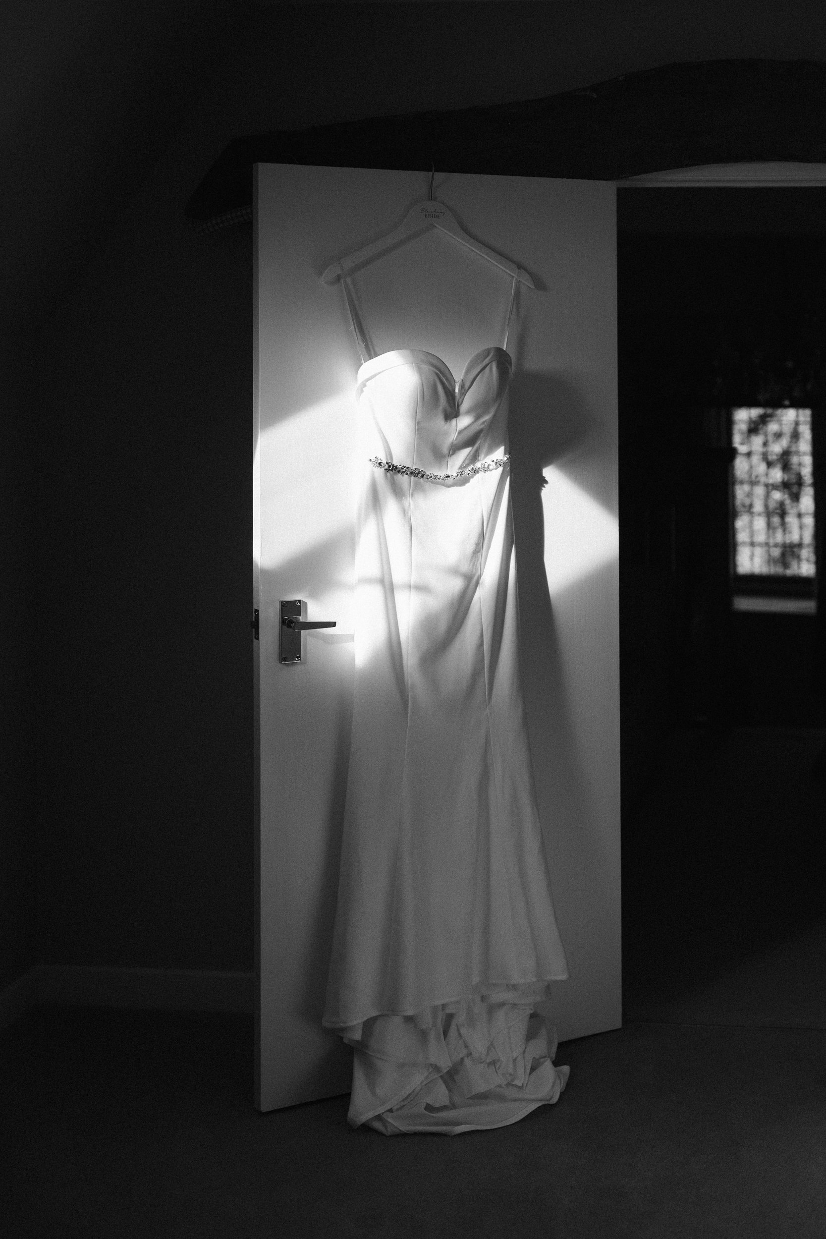 A wedding dress hanging on a hanger on a door, illuminated by a beam of light in a dimly lit room.