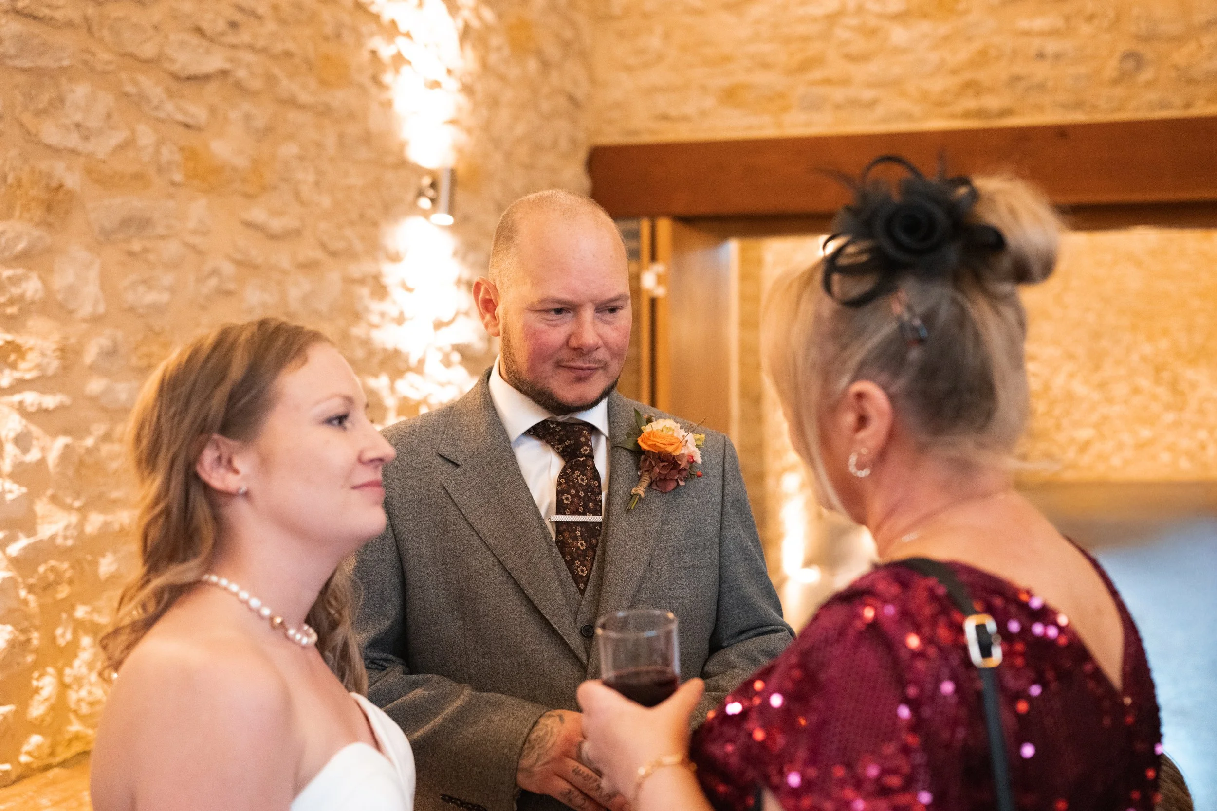 A bride and groom are standing together during their wedding ceremony while a woman with gray hair, wearing a red sequined dress and a black fascinator, holds a glass of red wine and speaks to them. The bride has blonde hair and wears a pearl necklac