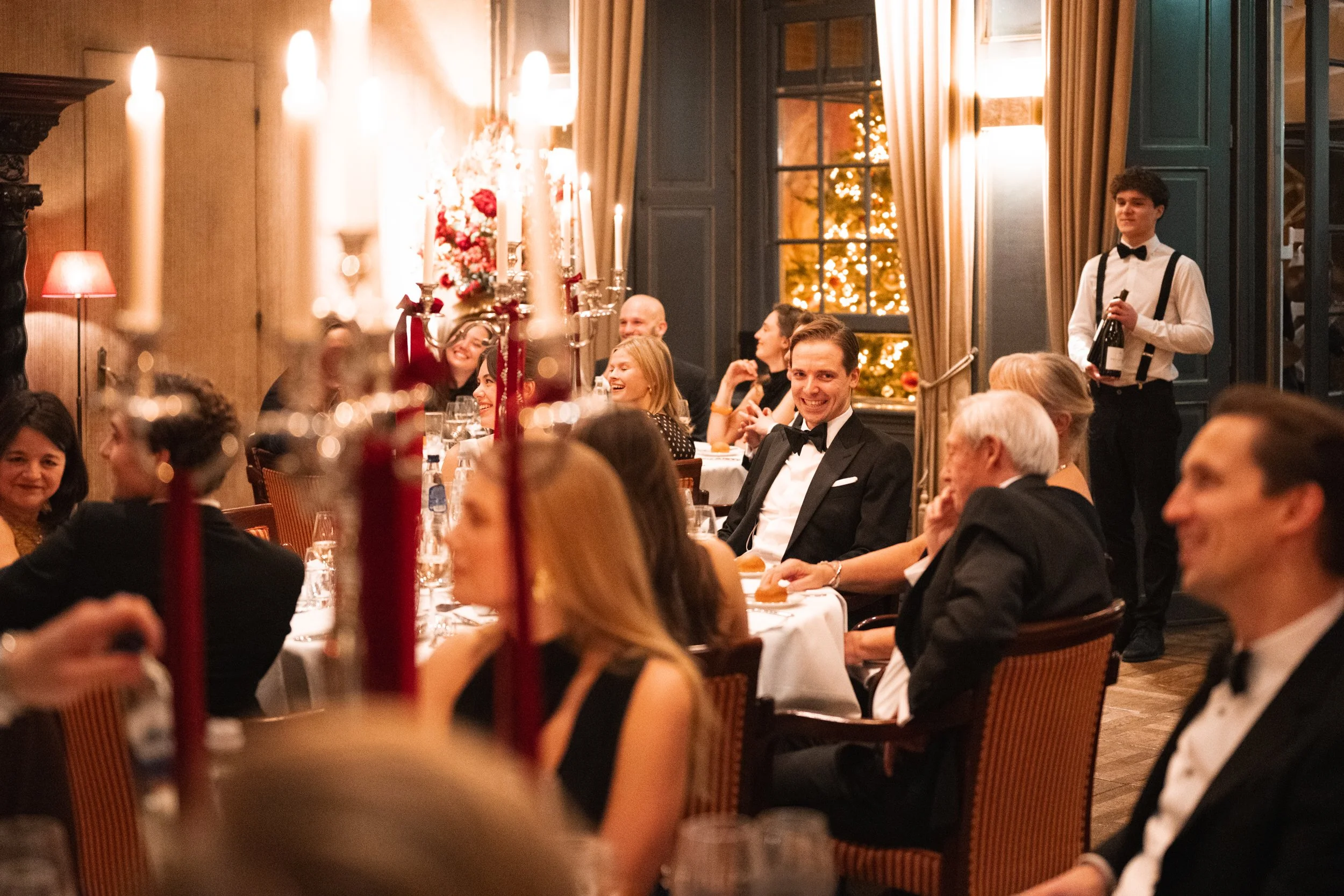 Guests in formal attire dining at a festive dinner party, with a man in a tuxedo smiling. The setting is decorated with candles, flowers, and Christmas trees, indicating a holiday celebration.