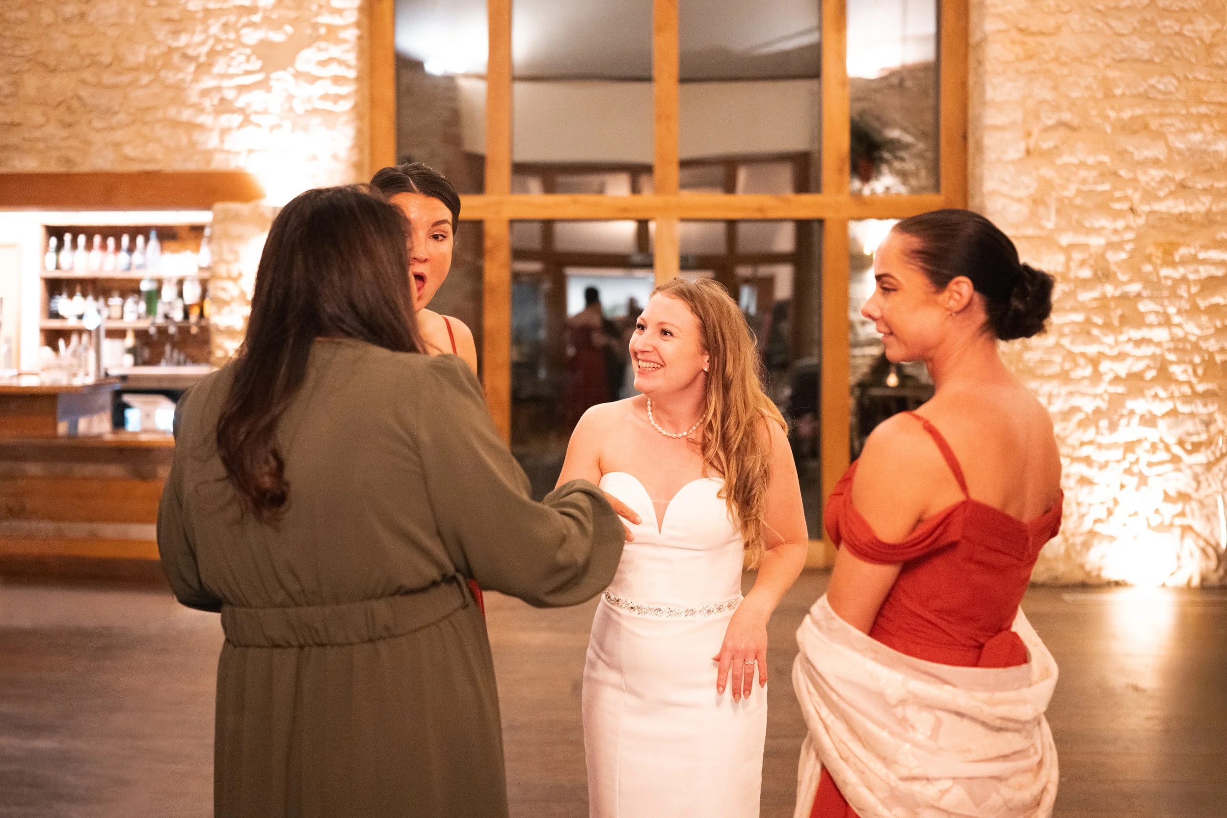 Four women are engaged in conversation at a social event inside a warmly lit venue with wooden beams and stone walls. One woman in a white dress is smiling, another in a red dress with a shawl, a third with dark hair in an olive dress, and the fourth
