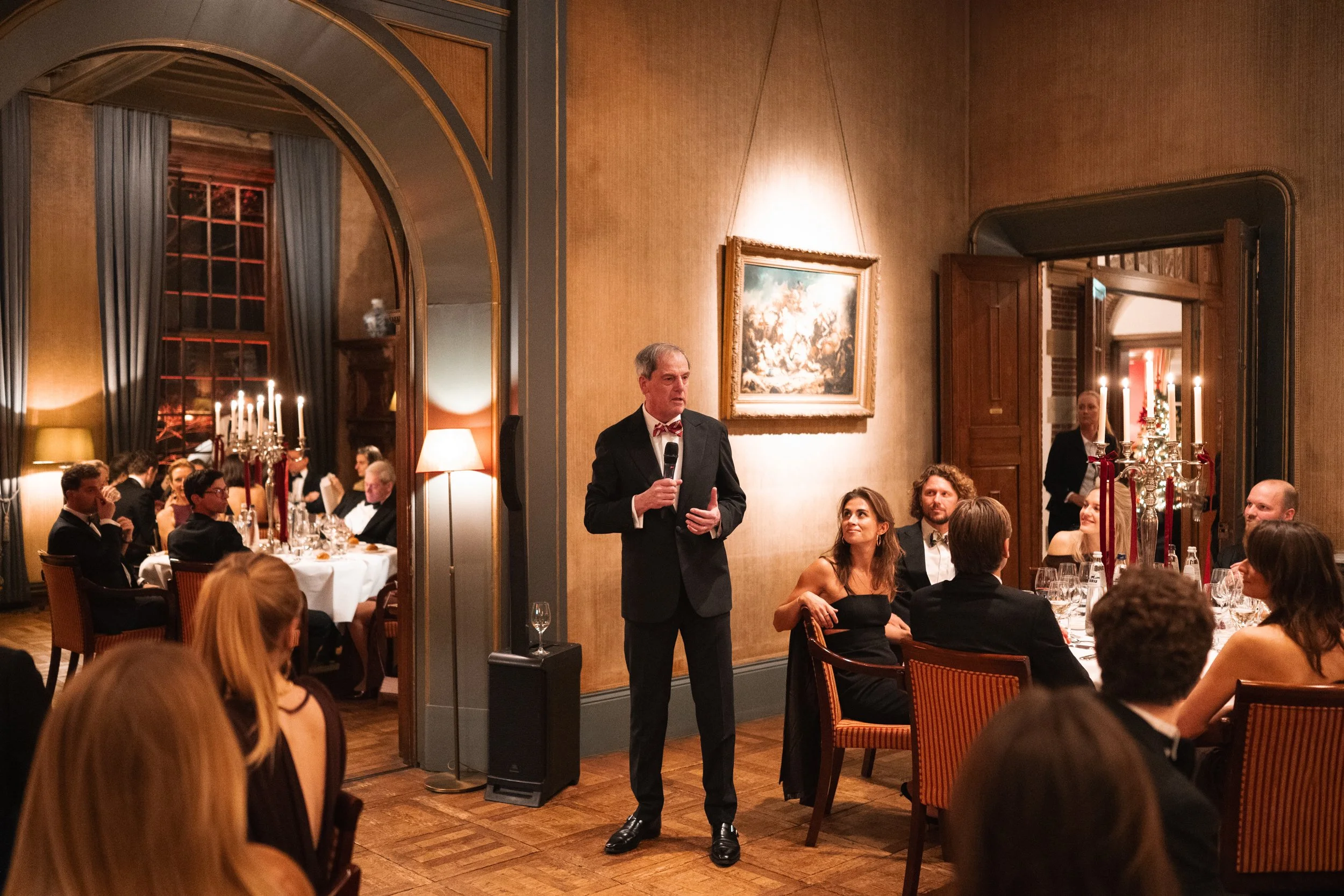 Man in tuxedo giving speech at a formal dinner event in an elegant dining room with guests seated at tables, candlelit chandeliers, and artwork on the walls.