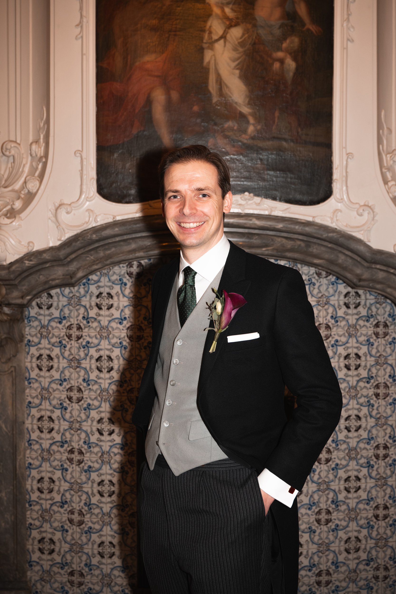 A man dressed in a black tuxedo with a gray vest, white shirt, and green tie, wearing a boutonnière, standing indoors in front of ornate wall decor and a large painting, smiling at the camera.