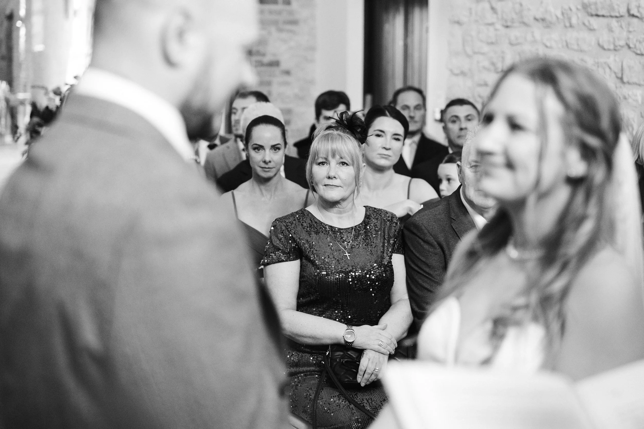 Black and white photo of a group of people at a formal event, focusing on an older woman in the center wearing a sequined dress and a cross necklace, with others around her listening attentively.