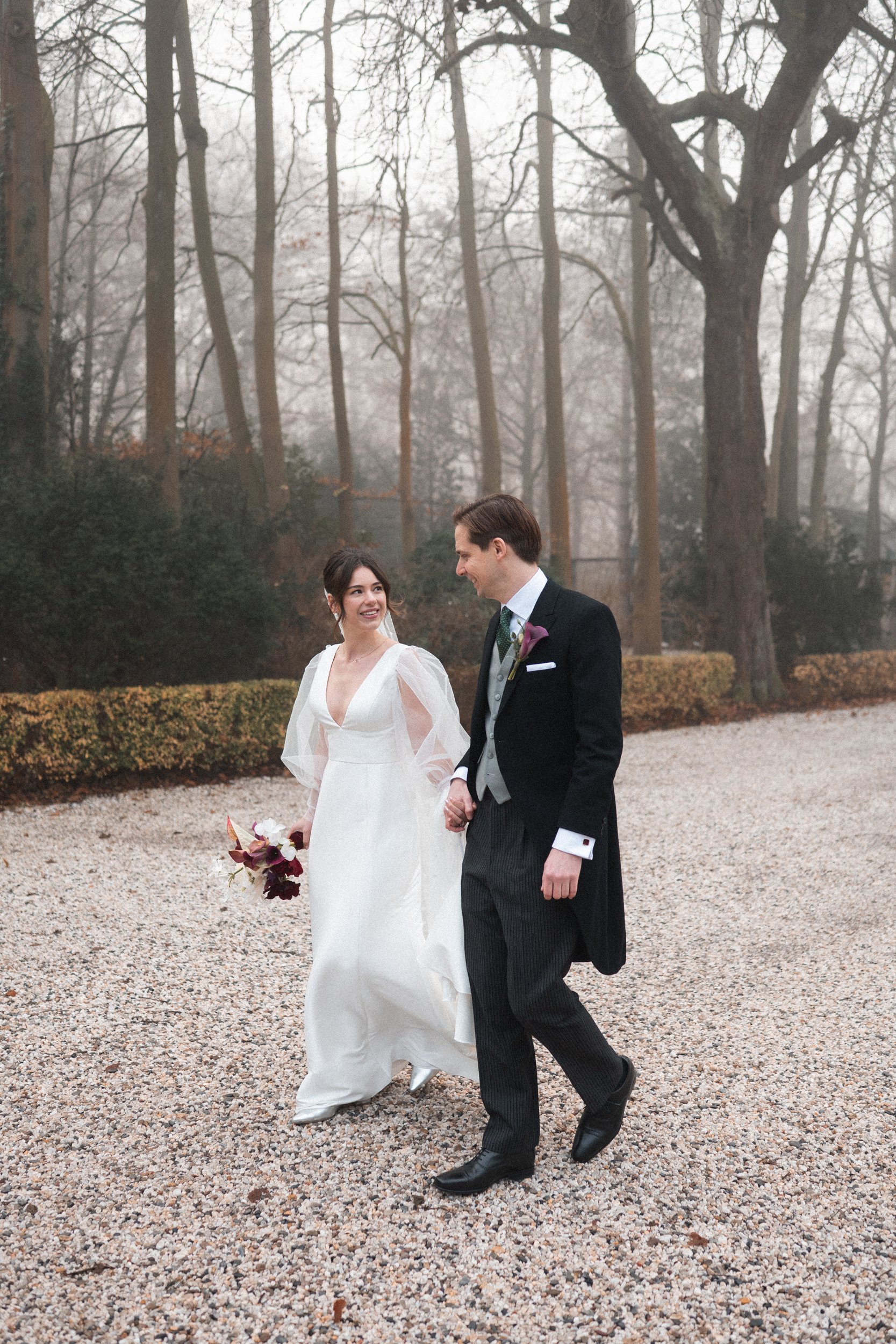 A newlywed couple walking outdoors on a gravel path in a wooded area with tall trees and fog, the bride holding a bouquet and wearing a white wedding gown, and the groom dressed in a black tuxedo with a green vest and purple boutonnière.