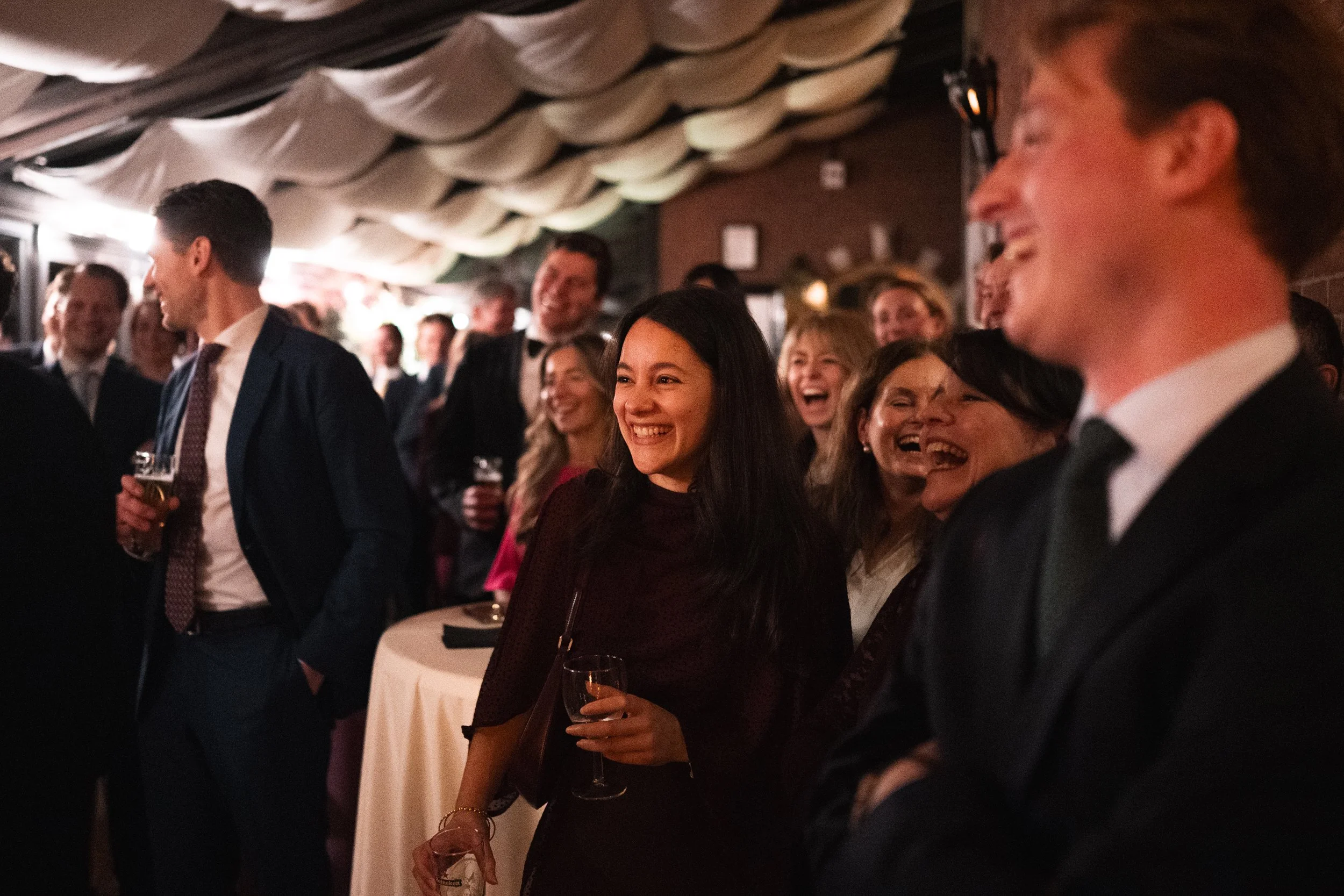 Group of people in formal attire enjoying a social event, smiling and laughing, in a dimly lit indoor setting.