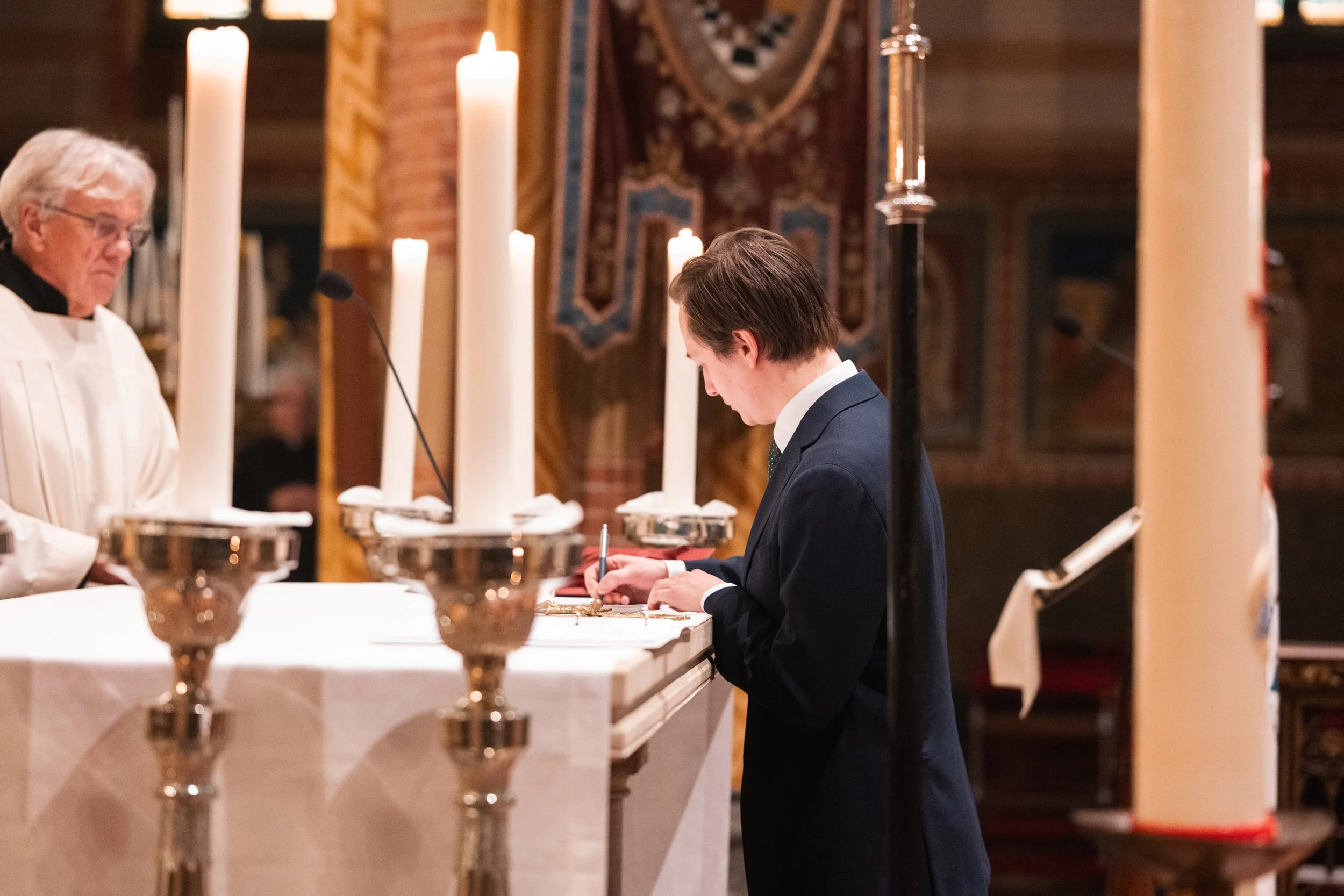 A young man in a navy suit is signing a book at an altar during a religious ceremony inside a church, with a priest standing nearby among lit candles and religious decorations in the background.