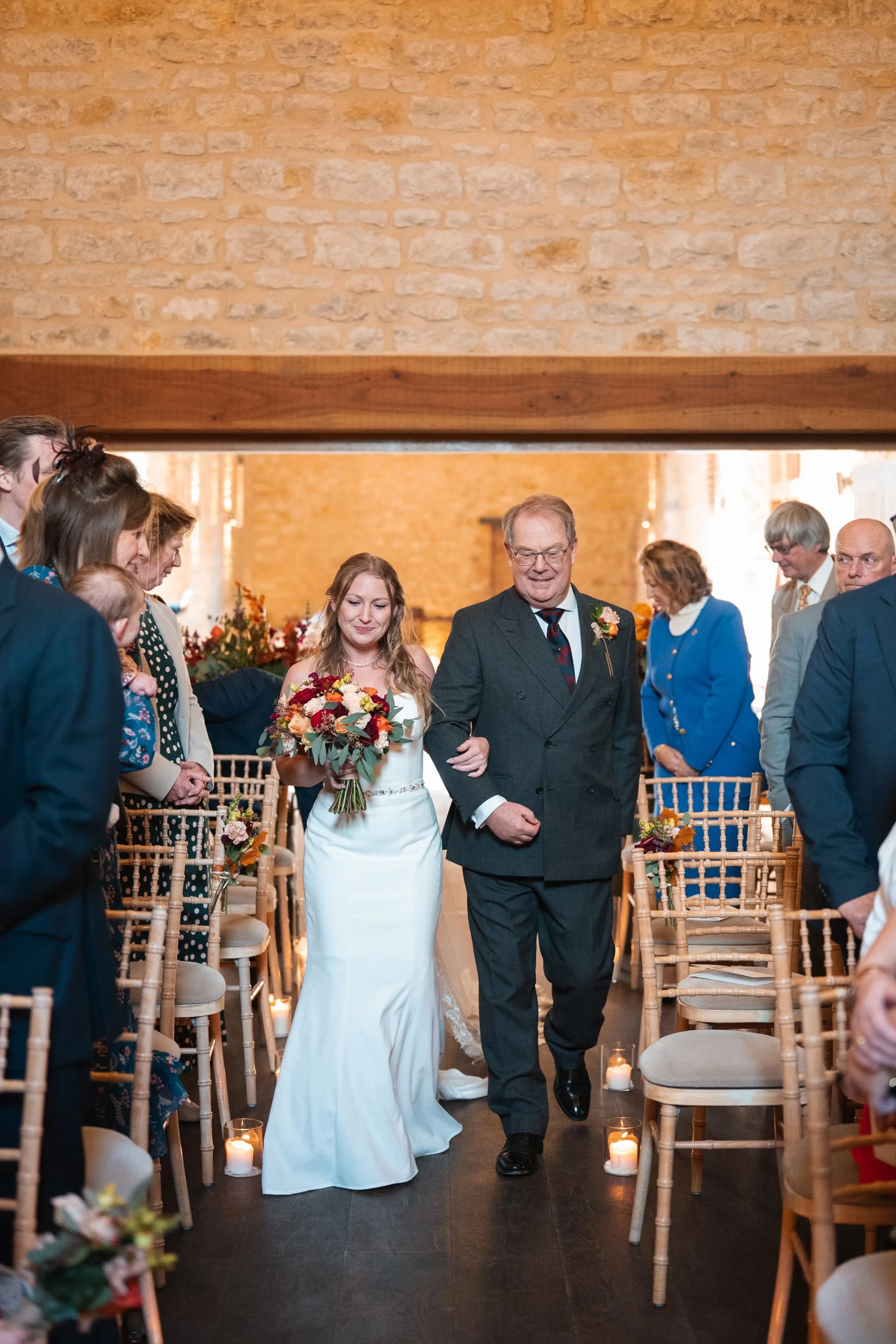 A bride walking down the aisle with her father during a wedding ceremony in a rustic venue with brick walls, surrounded by seated guests.