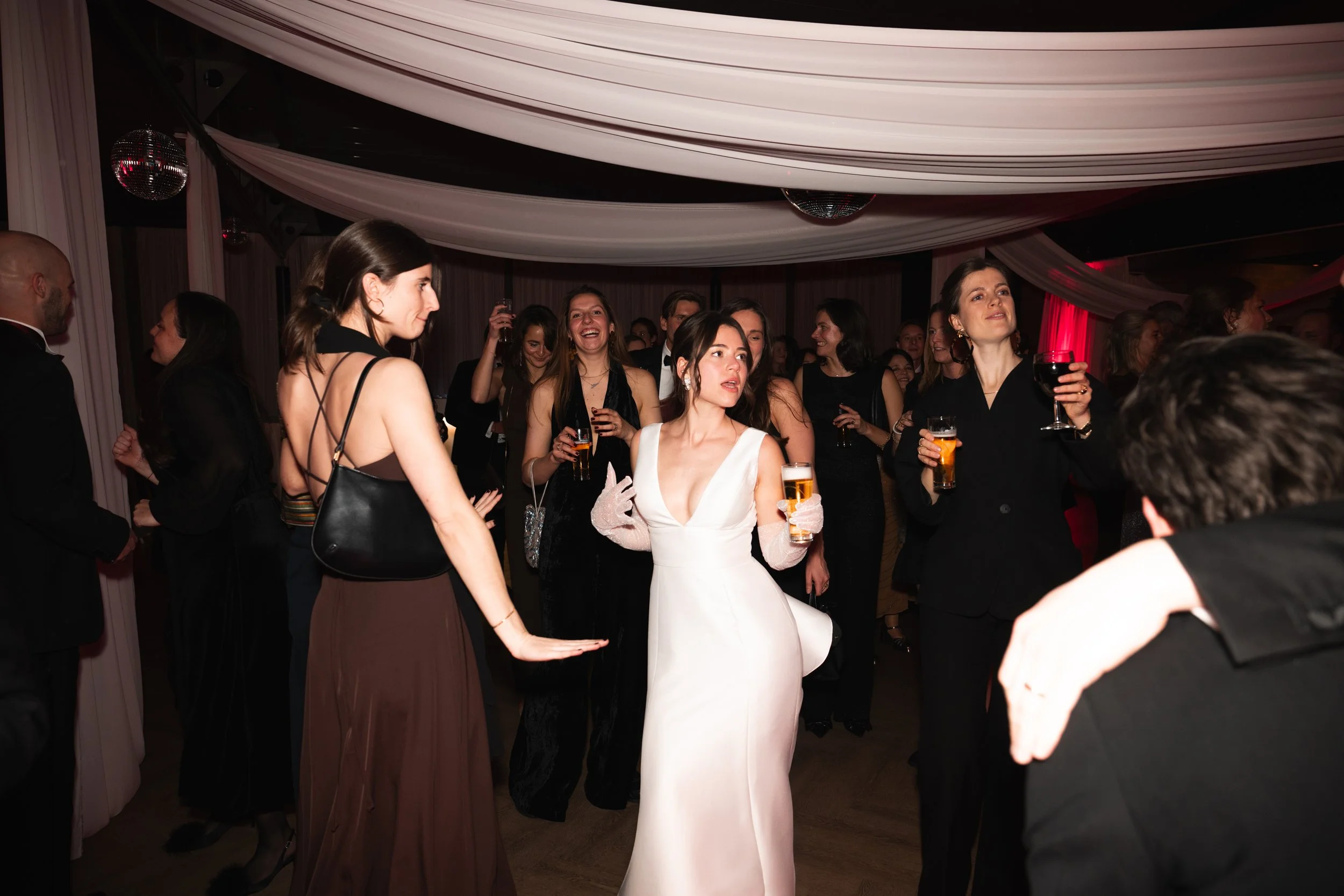 Women in formal attire at a social event, holding drinks, and engaging in conversation, with decorative drapes and disco balls overhead.