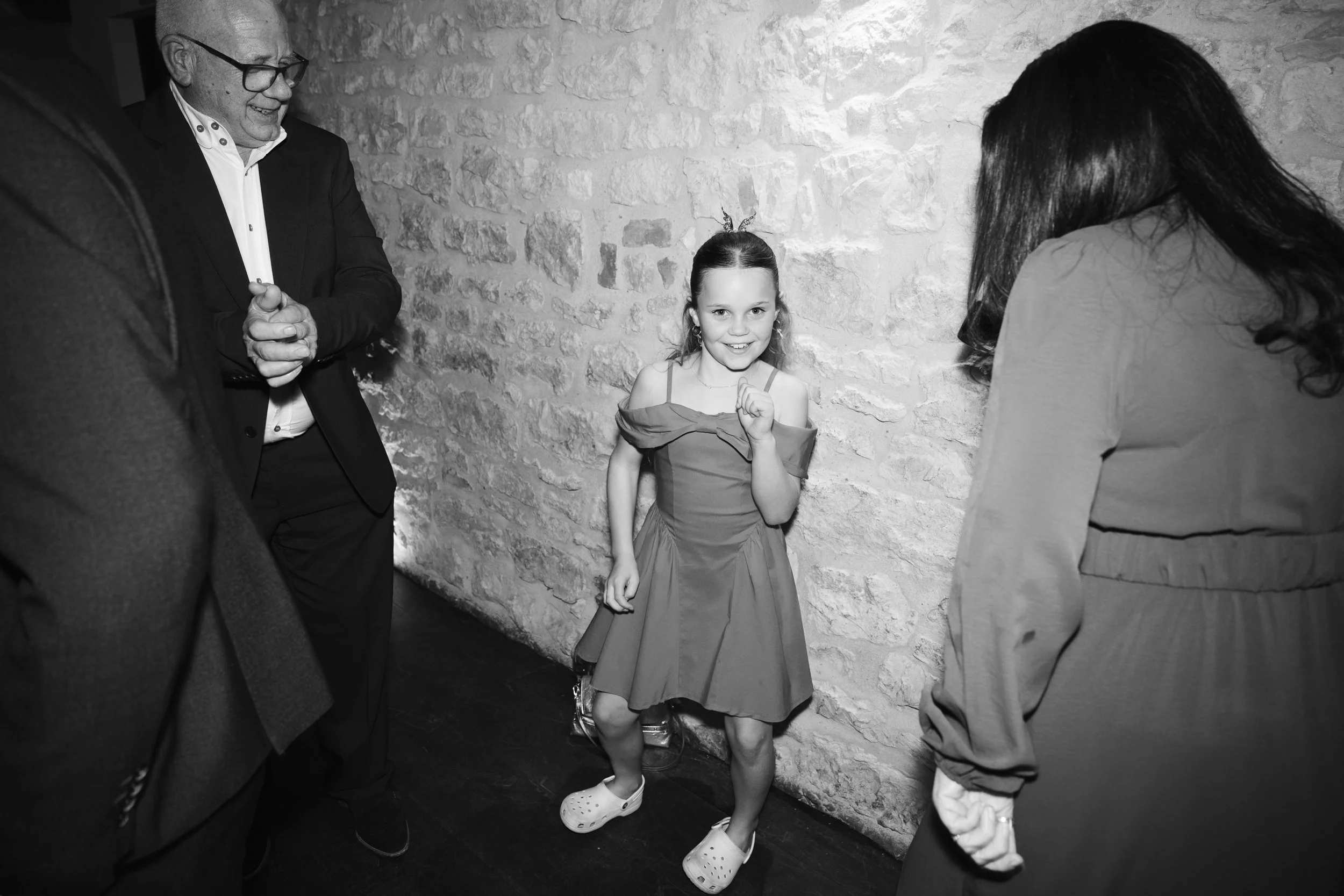 A young girl with a happy expression, wearing a dress and Crocs, standing against a stone wall and surrounded by adults at an indoor gathering.