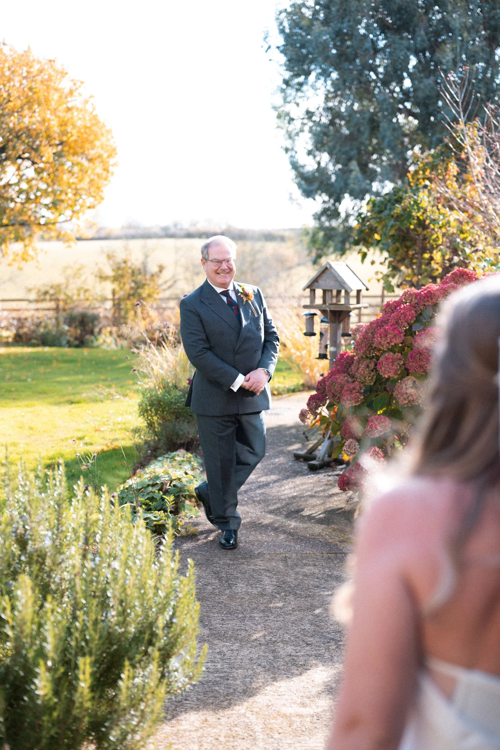 A man in a gray suit smiling and walking towards a woman with long, blonde hair during an outdoor event on a sunny day, with flowers and trees in the background.