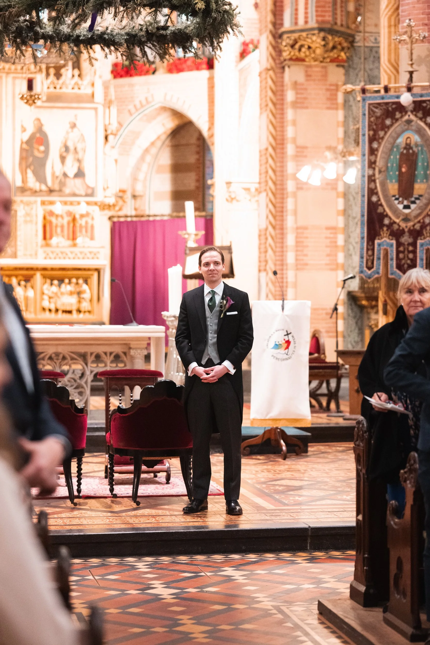 A young man dressed in a formal black suit with a gray vest and tie, standing inside a decorated church with ornate wooden furniture and religious icons on the walls. He appears to be a groom at a wedding, surrounded by other guests.