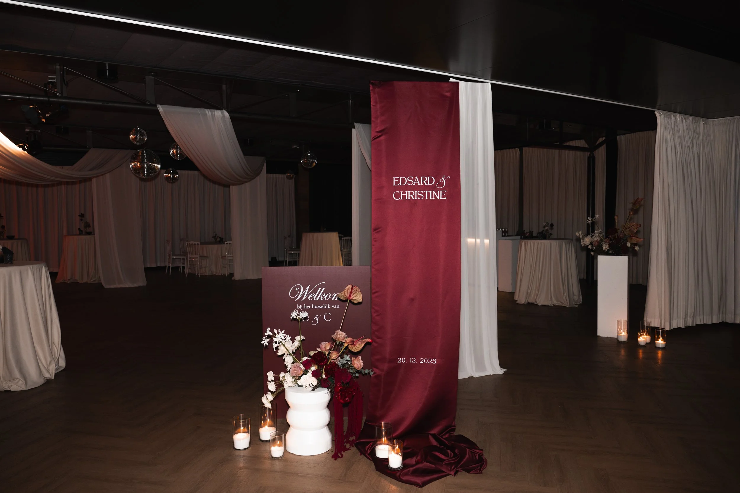 Wedding reception hall decorated with white draped fabric, flowers, candles, and a burgundy welcome sign with the names Edsard and Christine, and the date December 20, 2025.