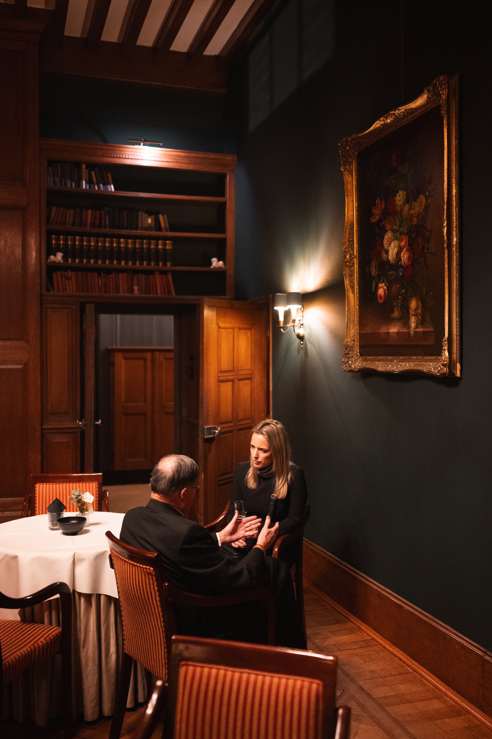 Two people sit at a table in a dimly lit, elegant room with dark walls, wooden accents, and a large framed painting of flowers. They are engaged in conversation, with one person gesturing and holding a glass.