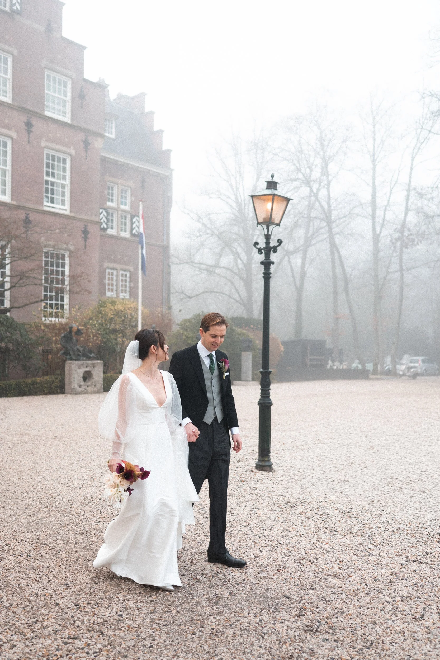 Bride and groom walking outside in wedding attire on a foggy day near a historic building and lamp post.