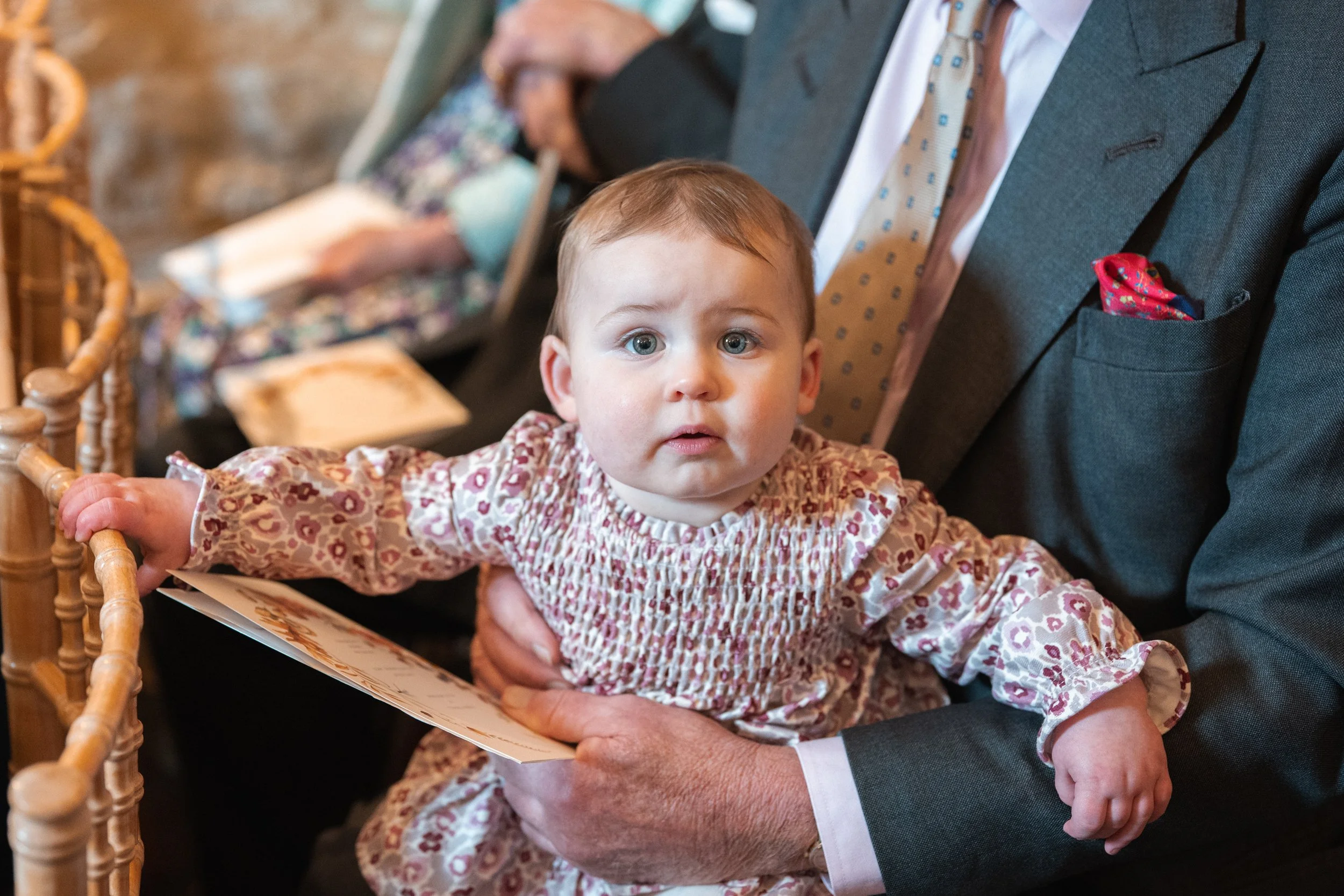 A baby girl with blue eyes and light brown hair being held on a man's lap, wearing a patterned dress in a formal setting with other people and wooden chairs in the background.