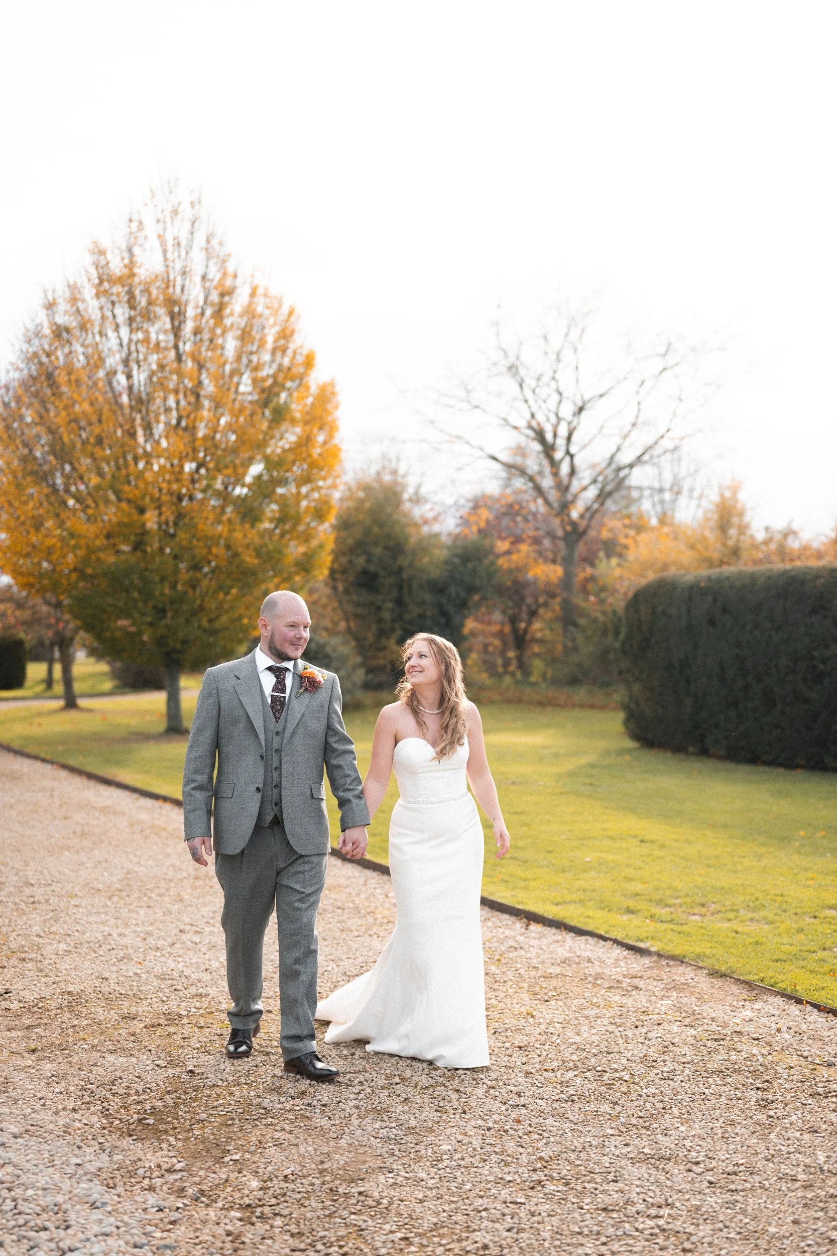 A newlywed couple holding hands and walking on a gravel path in a park surrounded by autumn trees, with the bride wearing a white strapless wedding gown and the groom in a gray suit.
