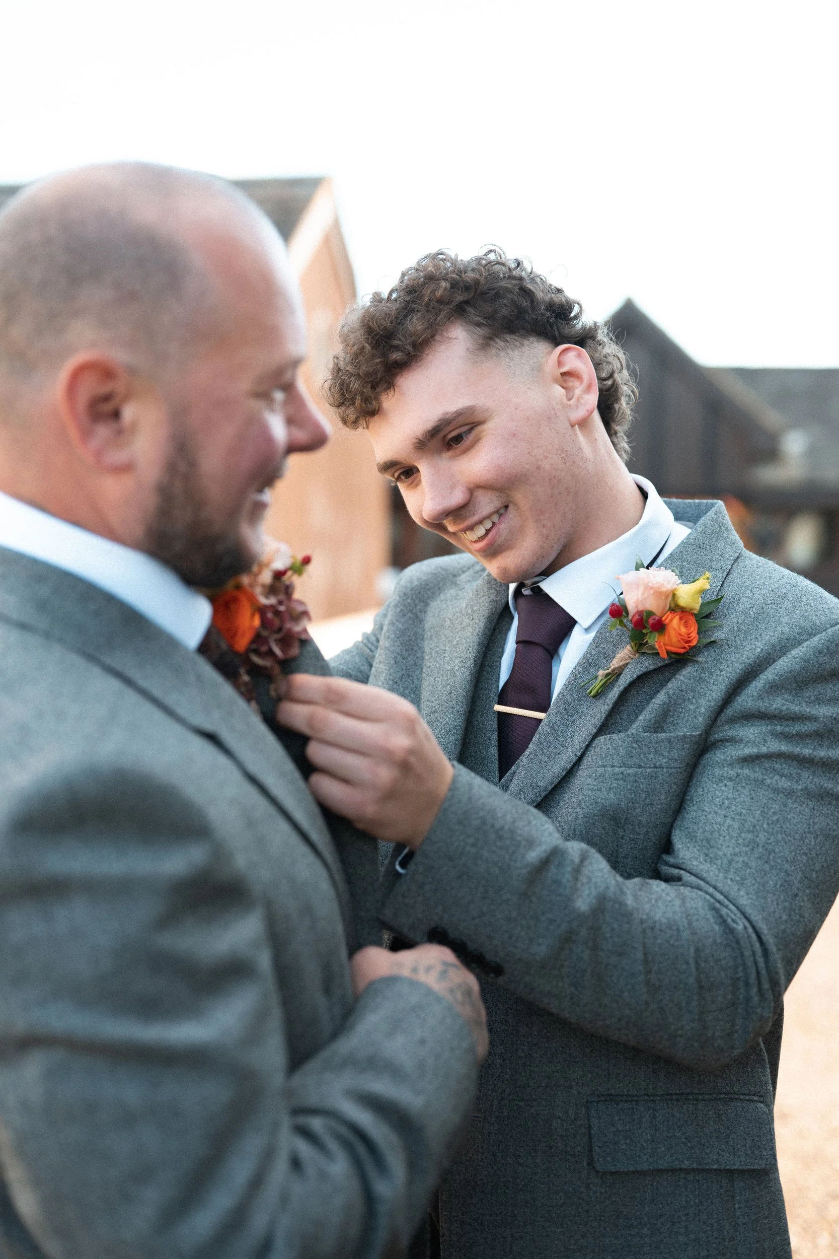 Two men in gray suits exchanging wedding vows outside, one fixing the other's lapel, both smiling, with a blurred background of houses.