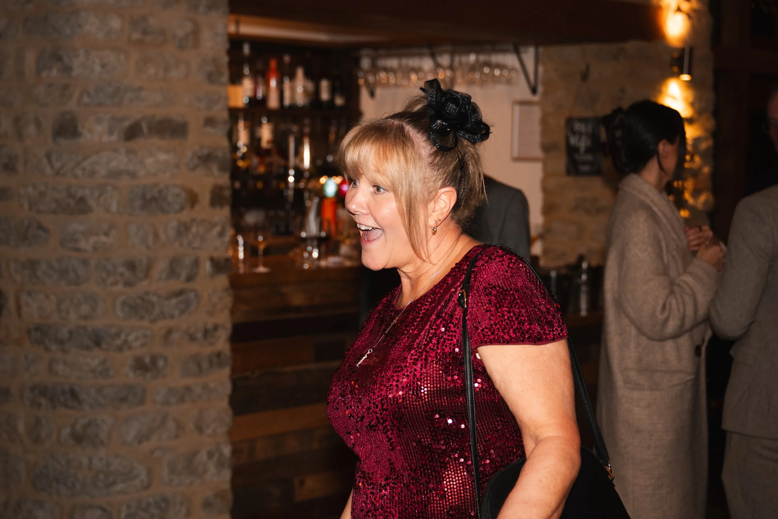 A woman laughing and talking at a social gathering in a bar, wearing a sequined red dress and a black hair accessory, with a bar and other people in the background.