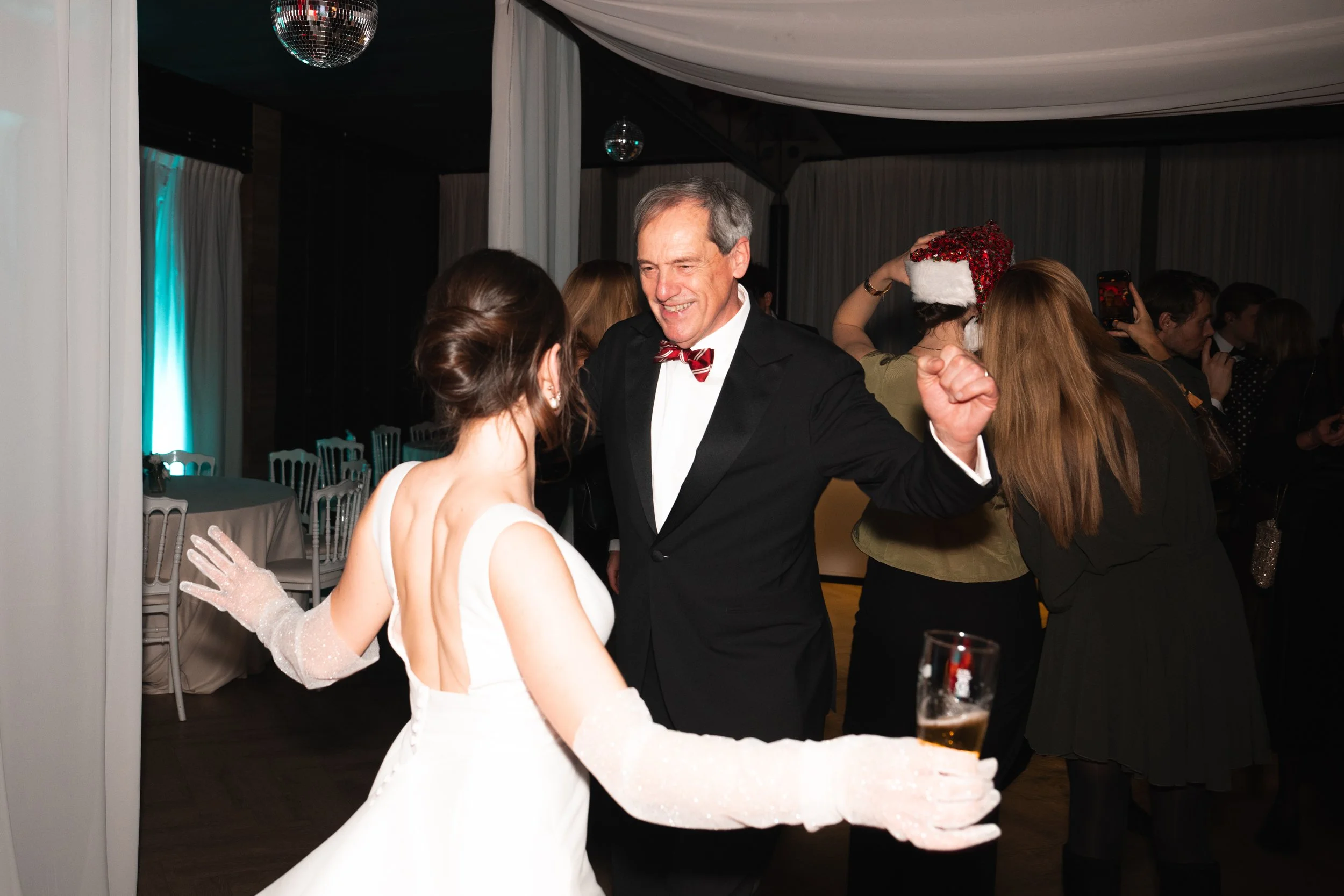 A wedding reception with the bride in a white gown and veil dancing with an older man in a tuxedo with a red bow tie. People in the background, some wearing Christmas hats, are dancing and taking photos. Disco balls hang from the ceiling.