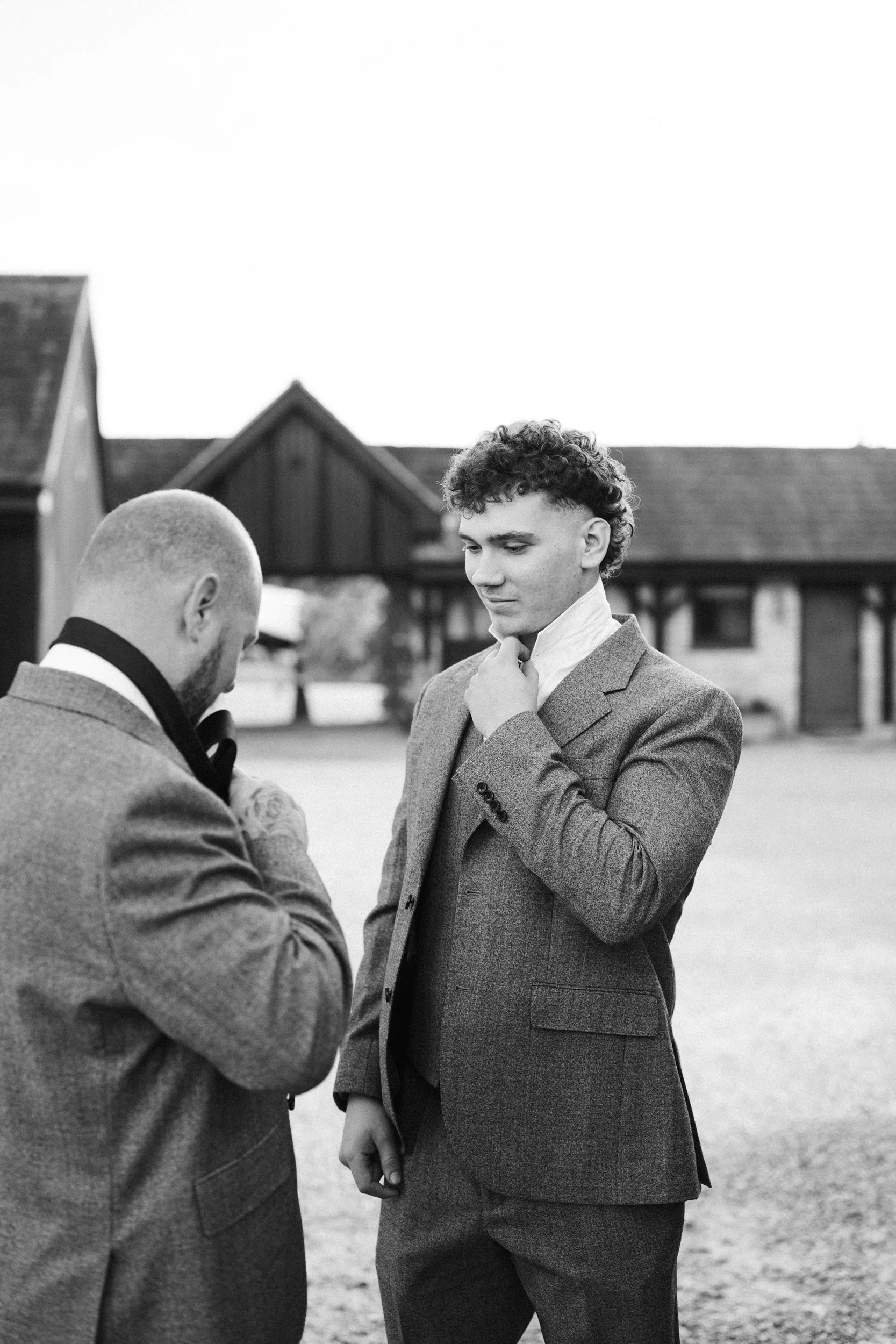 Two men in suits with one adjusting his jacket while the other looks on, outdoors in front of a barn-style building, in black and white.