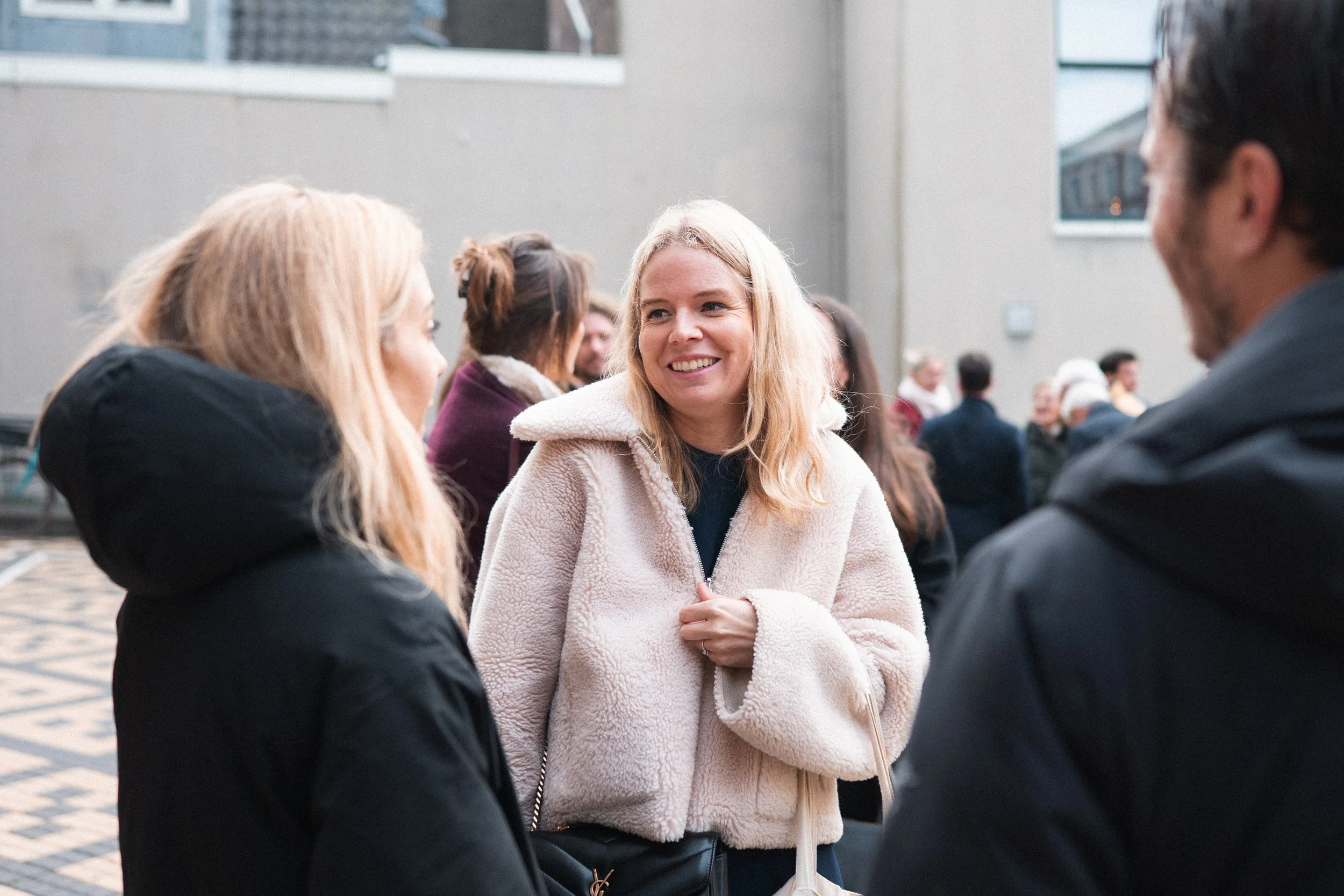 Group of people outdoors, talking and smiling, with a woman in a beige coat in the center.