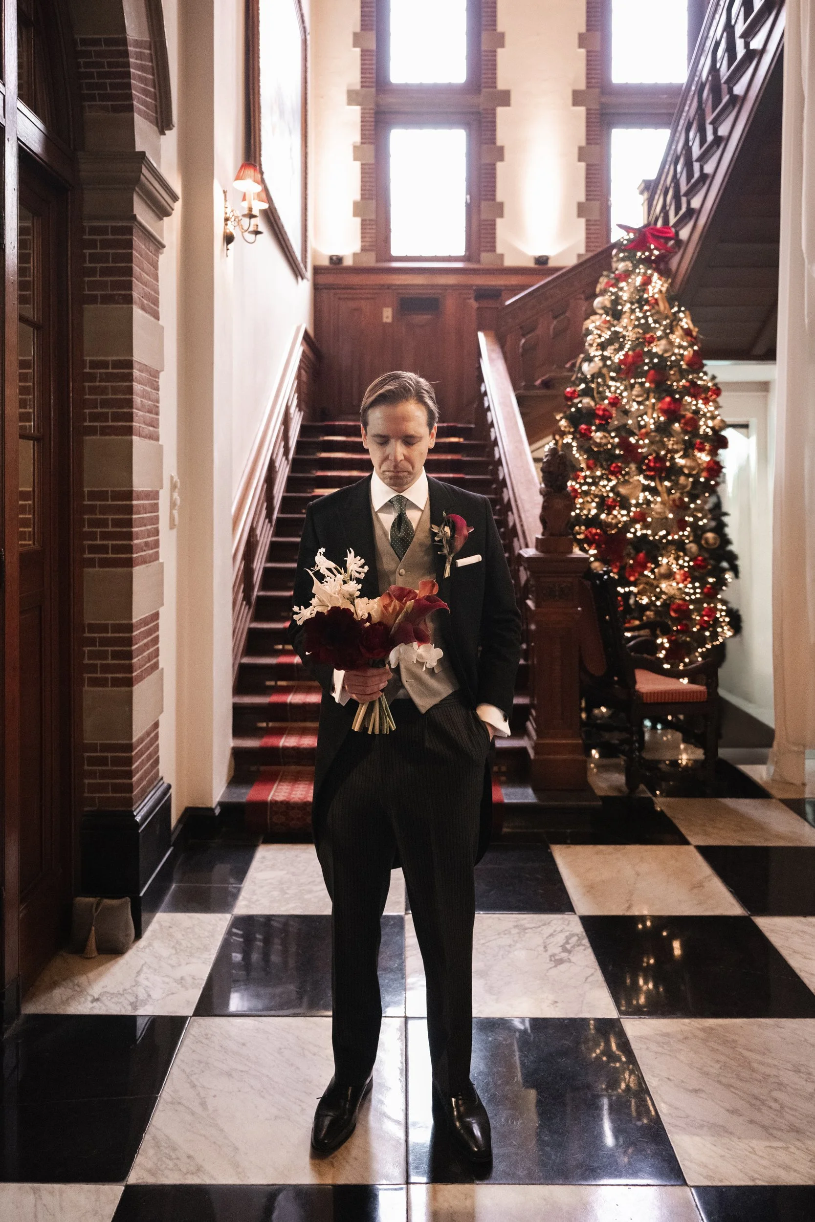 Man in formal attire holding a bouquet, standing in a decorated, elegant hallway with a Christmas tree, staircase, and checkered floor.