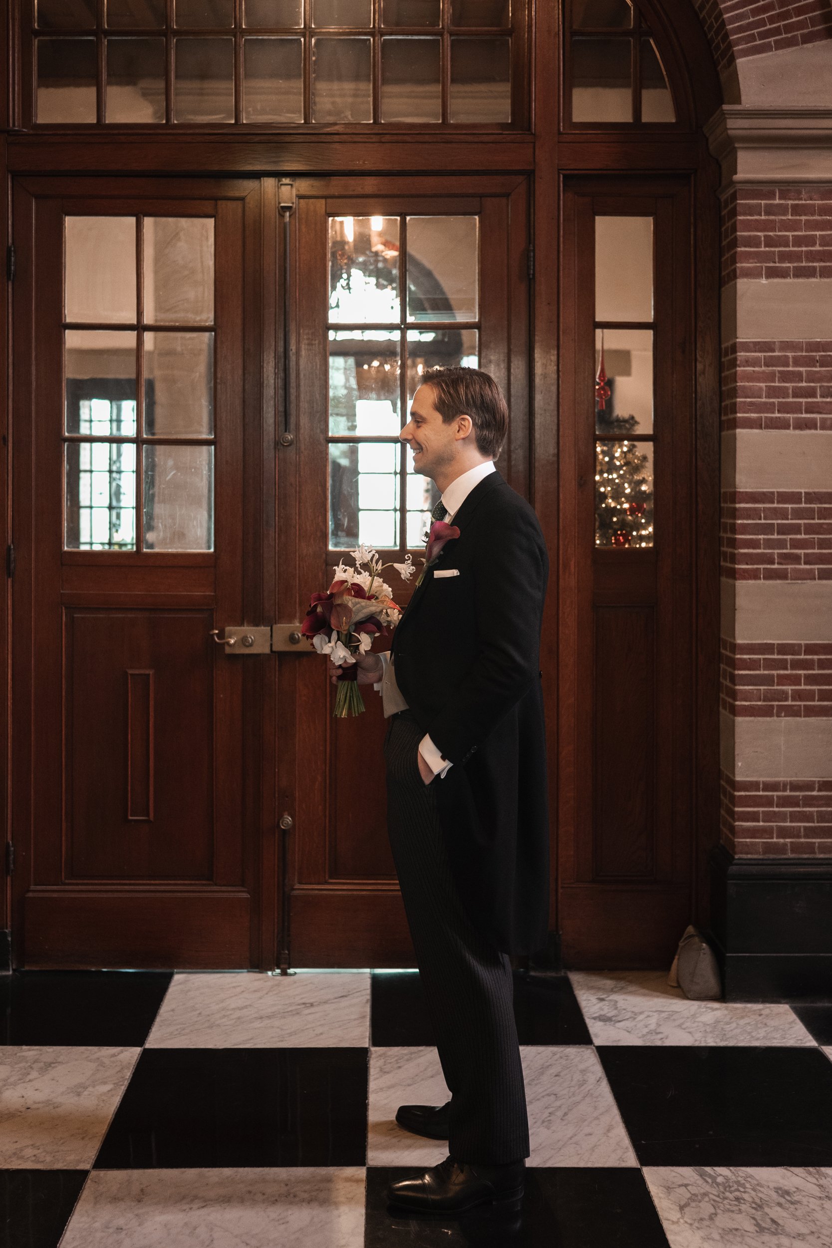 A man in a tuxedo holding a flower bouquet, standing inside a building with Victorian-style wooden doors and a Christmas tree visible through a window in the background.