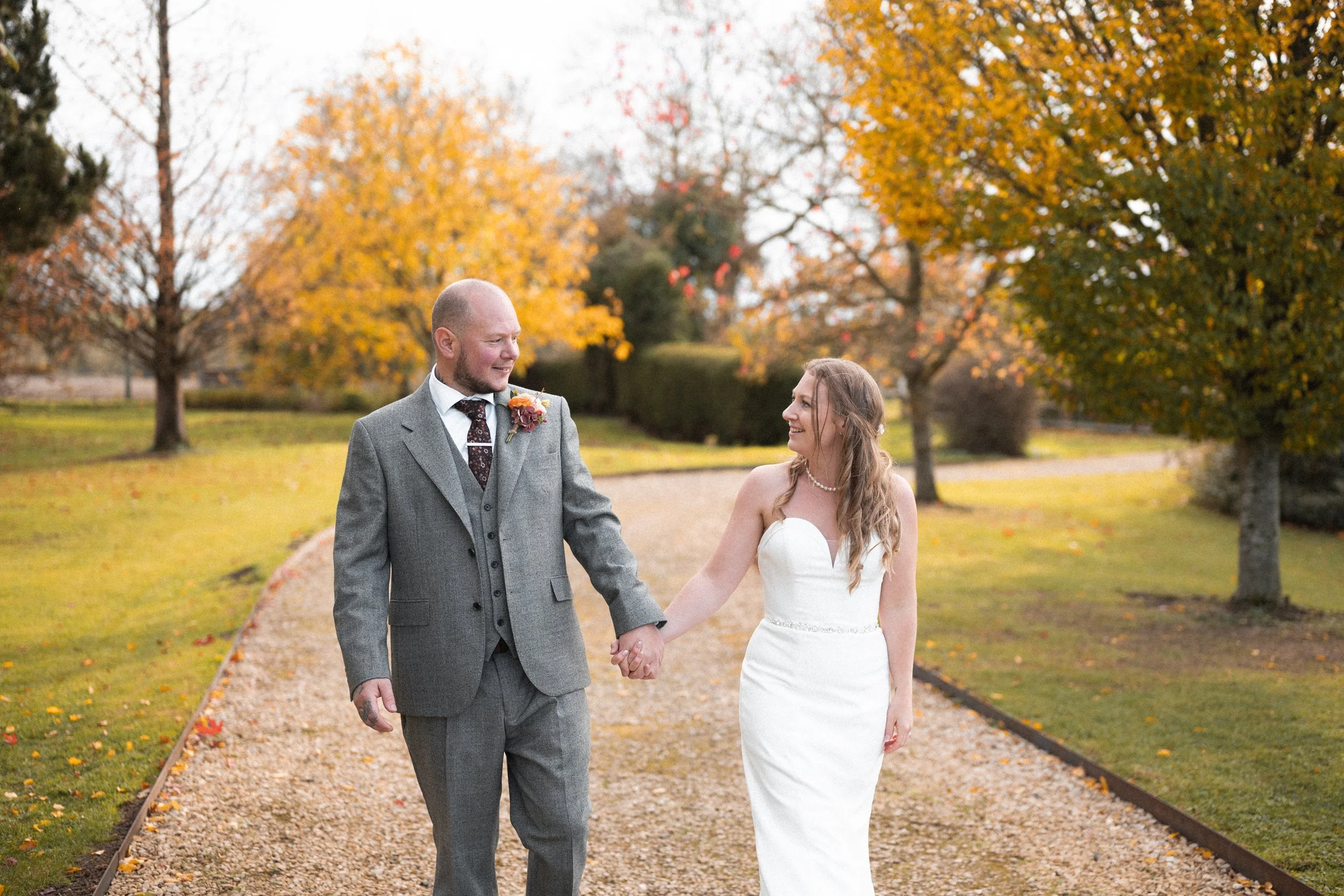 A bride and groom holding hands and walking in a park with autumn trees.