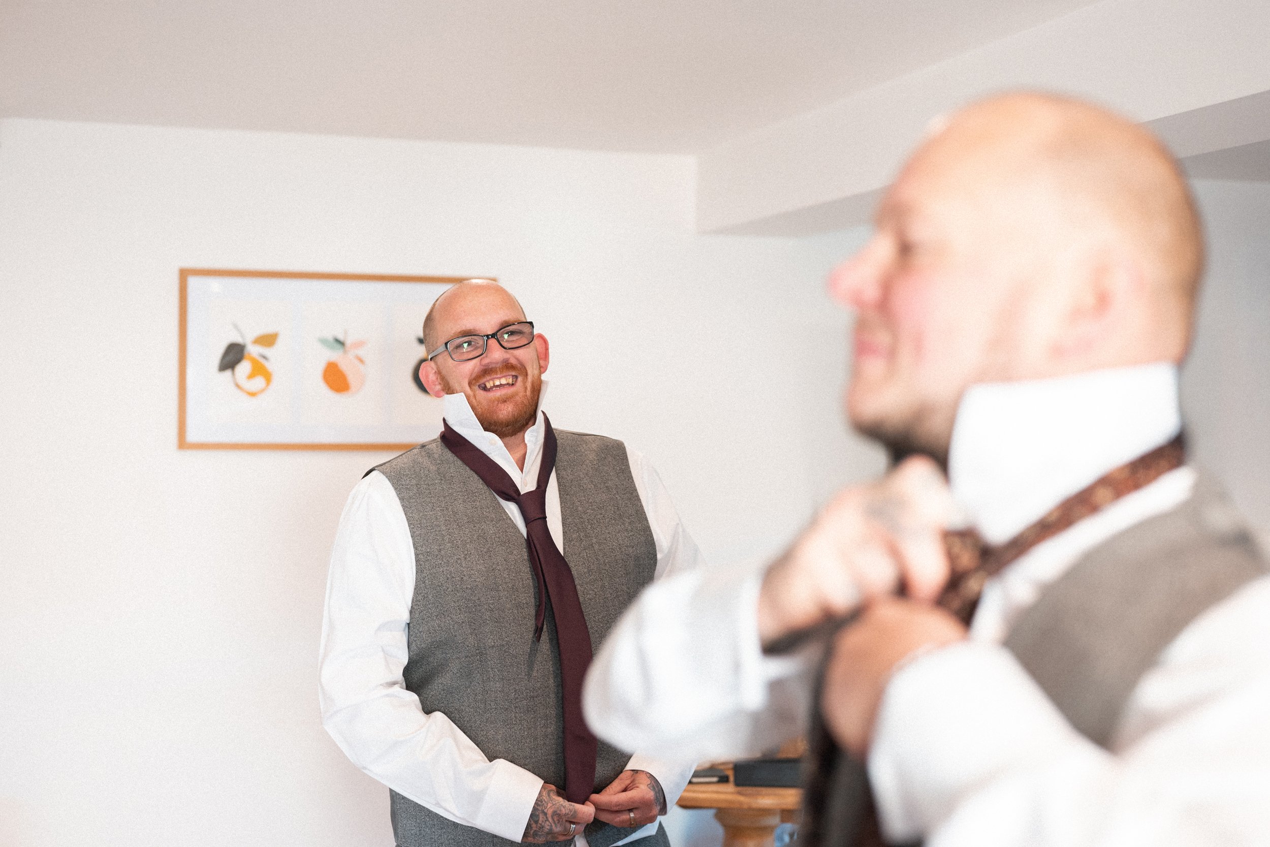Two men in formal attire holding ties, smiling and preparing for a formal event in a bright room with a fruit-themed framed picture on the wall behind them.