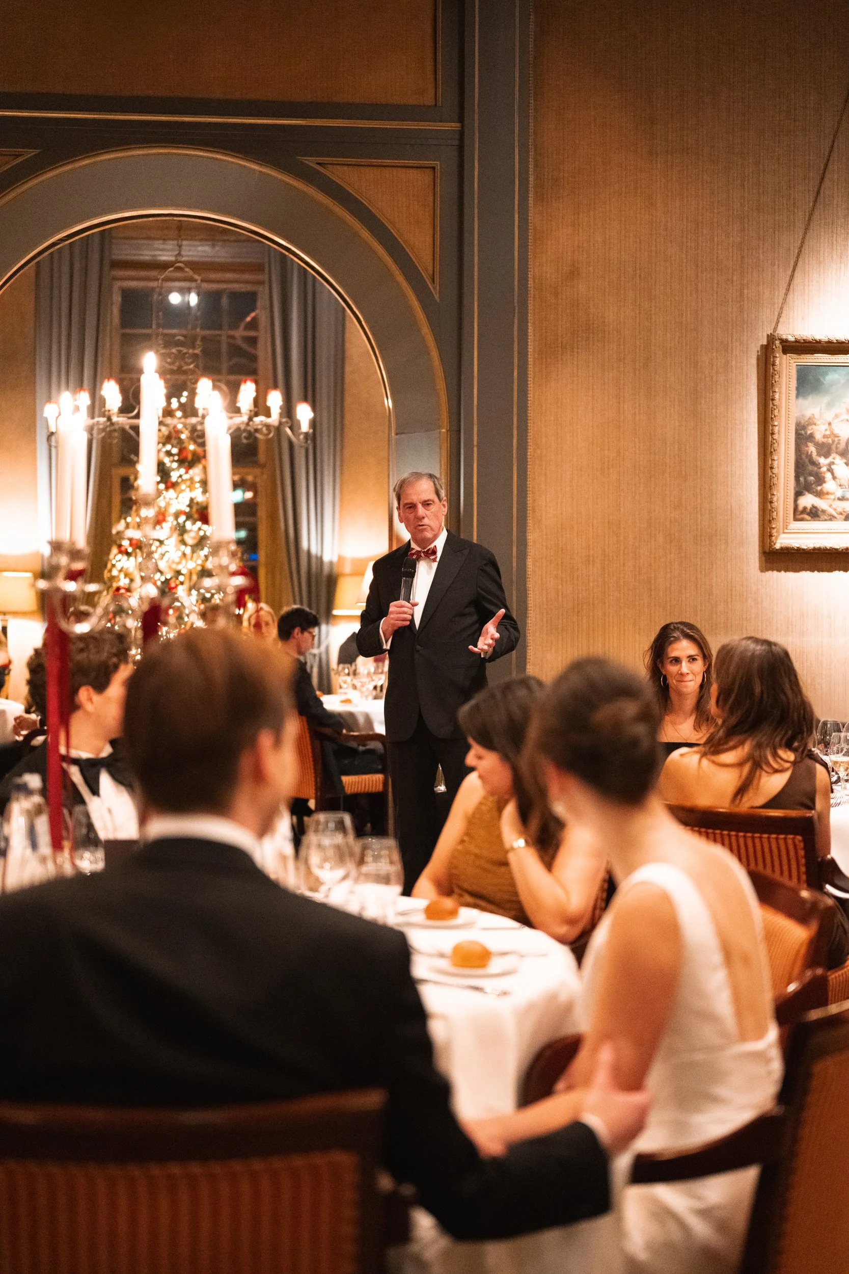 A man in a tuxedo giving a speech in a decorated holiday dinner setting, with a Christmas tree in the background.