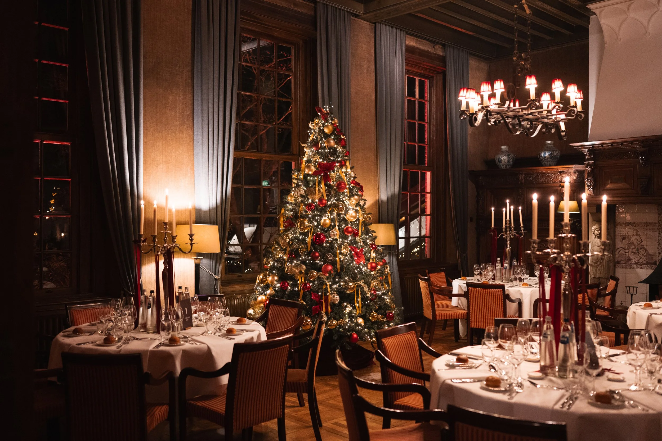 A decorated Christmas tree in a dimly lit dining room with tables set for a festive dinner, tall candles, ornate curtains, and chandeliers.