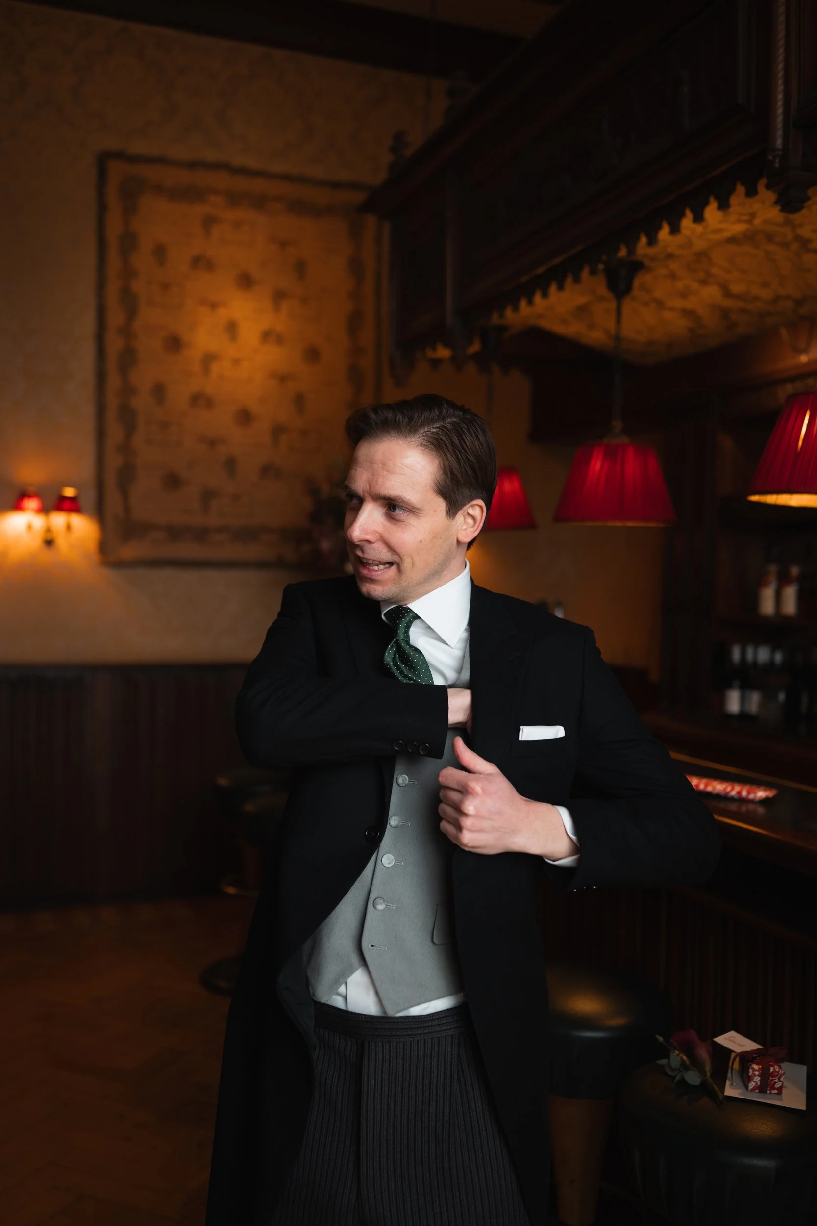 A man in formal attire adjusting his jacket in a warmly lit bar or lounge with red lamps and wooden decor.