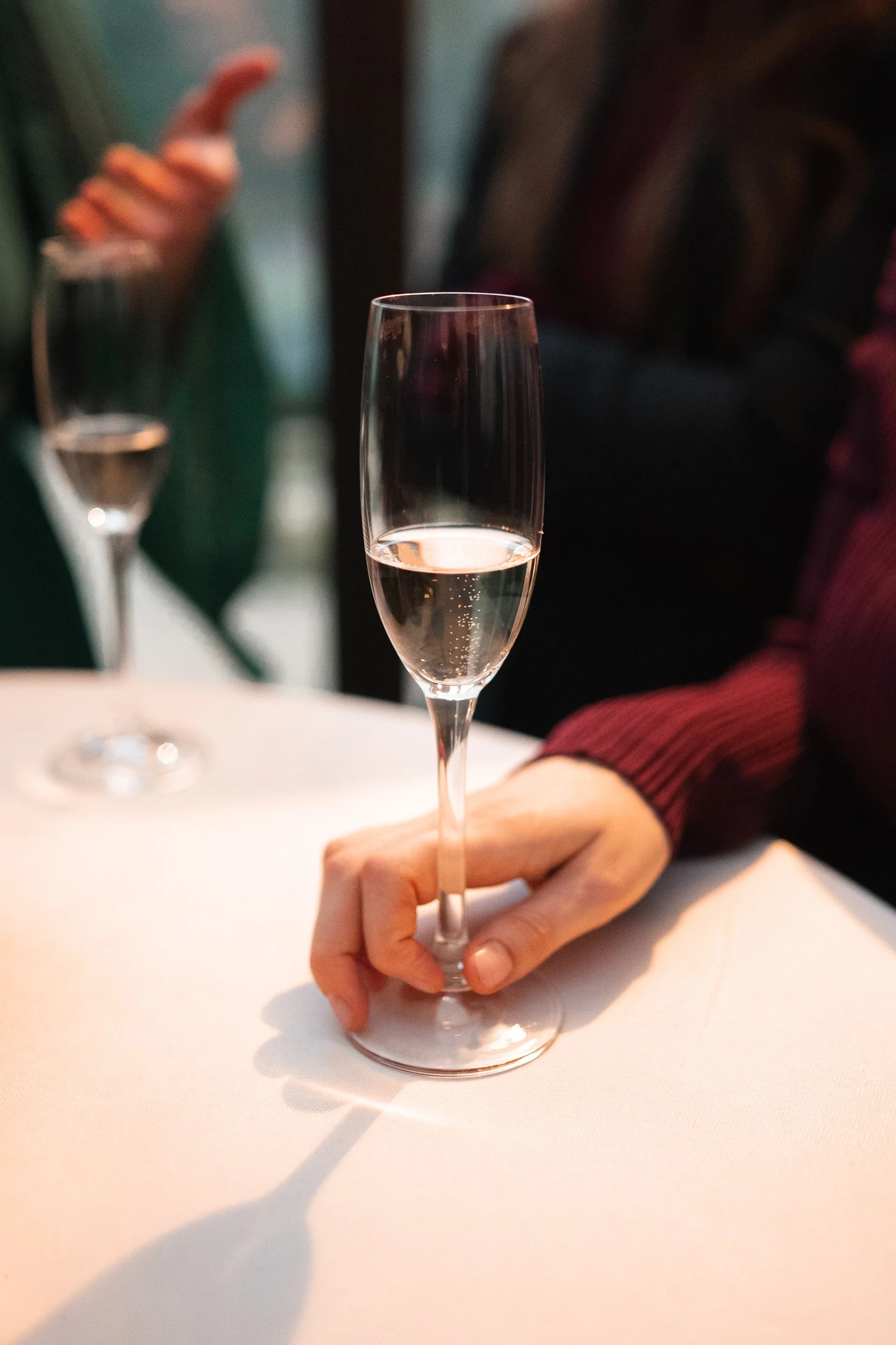 Person holding a champagne flute filled with sparkling wine on a white tablecloth with another partially visible champagne glass in the background.