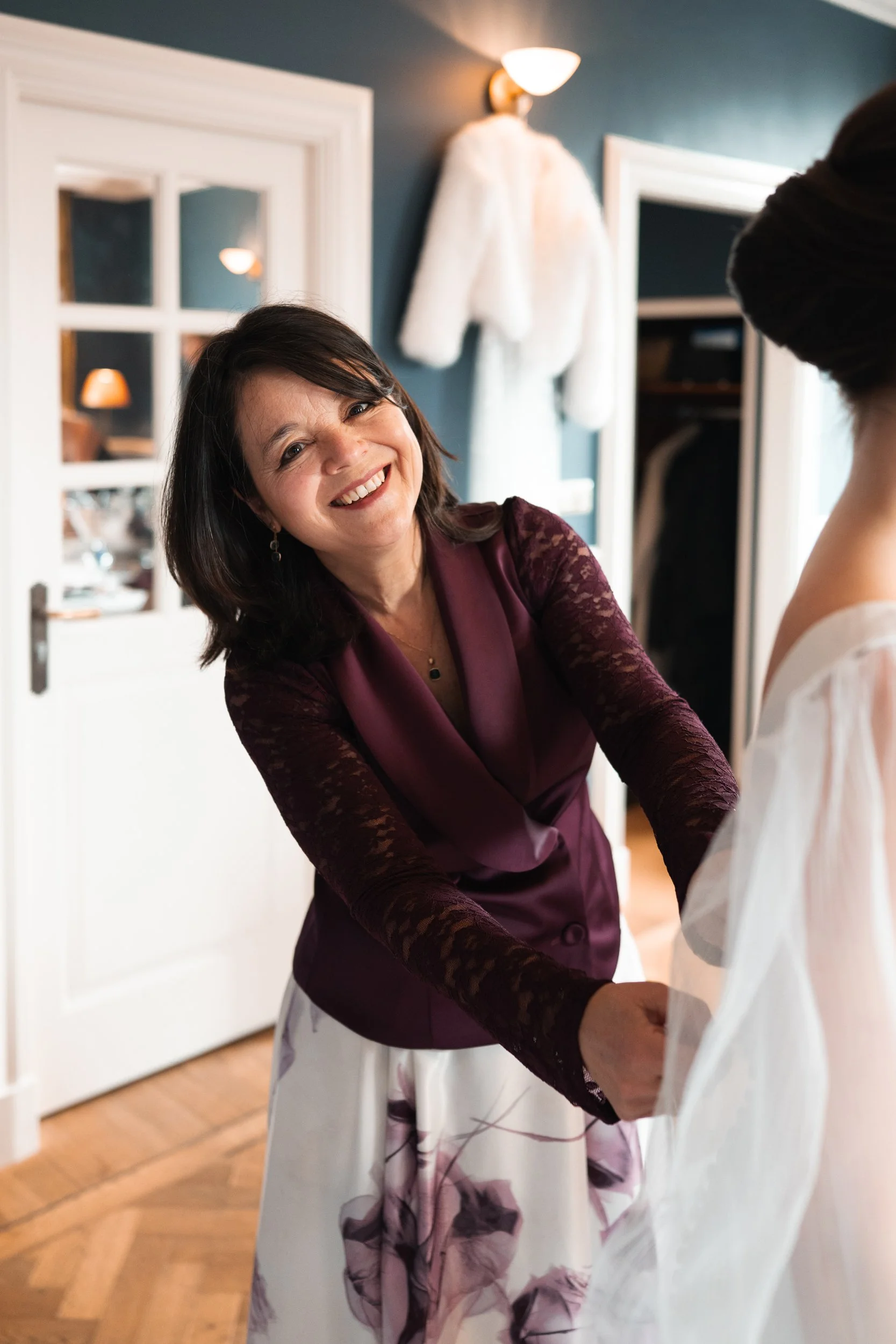 A woman in a purple jacket and floral skirt smiling as she helps a bride with her wedding dress.