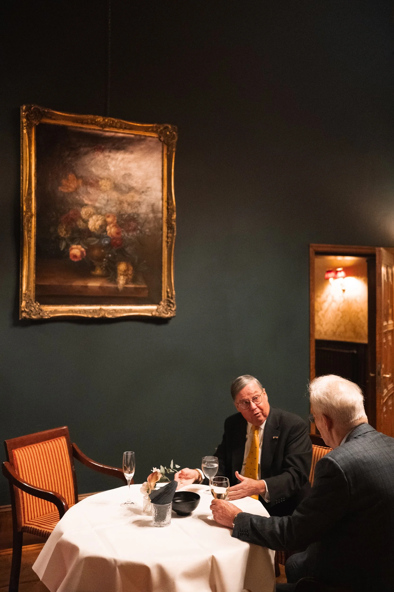 Two elderly men in suits having a conversation at a round dinner table with a white tablecloth, wine glasses, a black bowl, and a flower arrangement. They are in a dimly lit room with dark green walls and framed paintings, including a floral still li