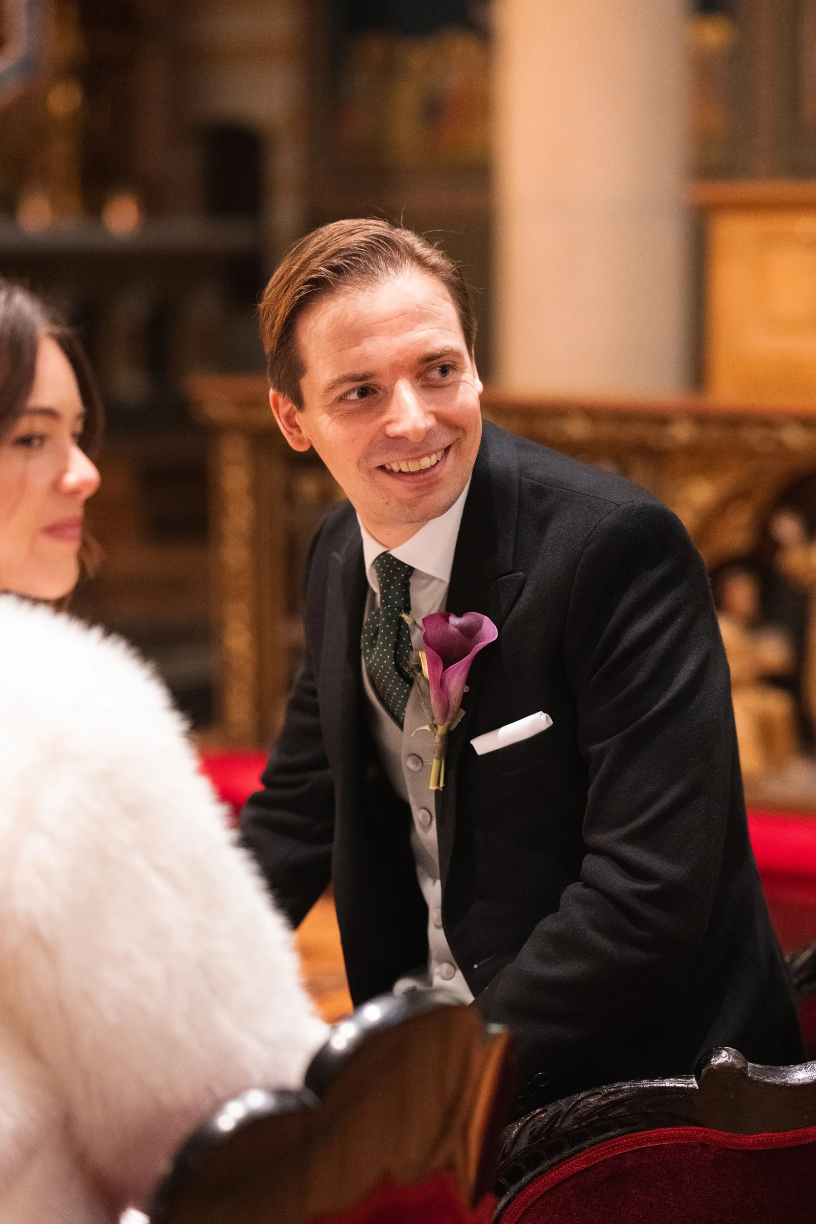 A man in a tuxedo with a purple calla lily boutonniere, smiling and leaning forward in a formal setting.