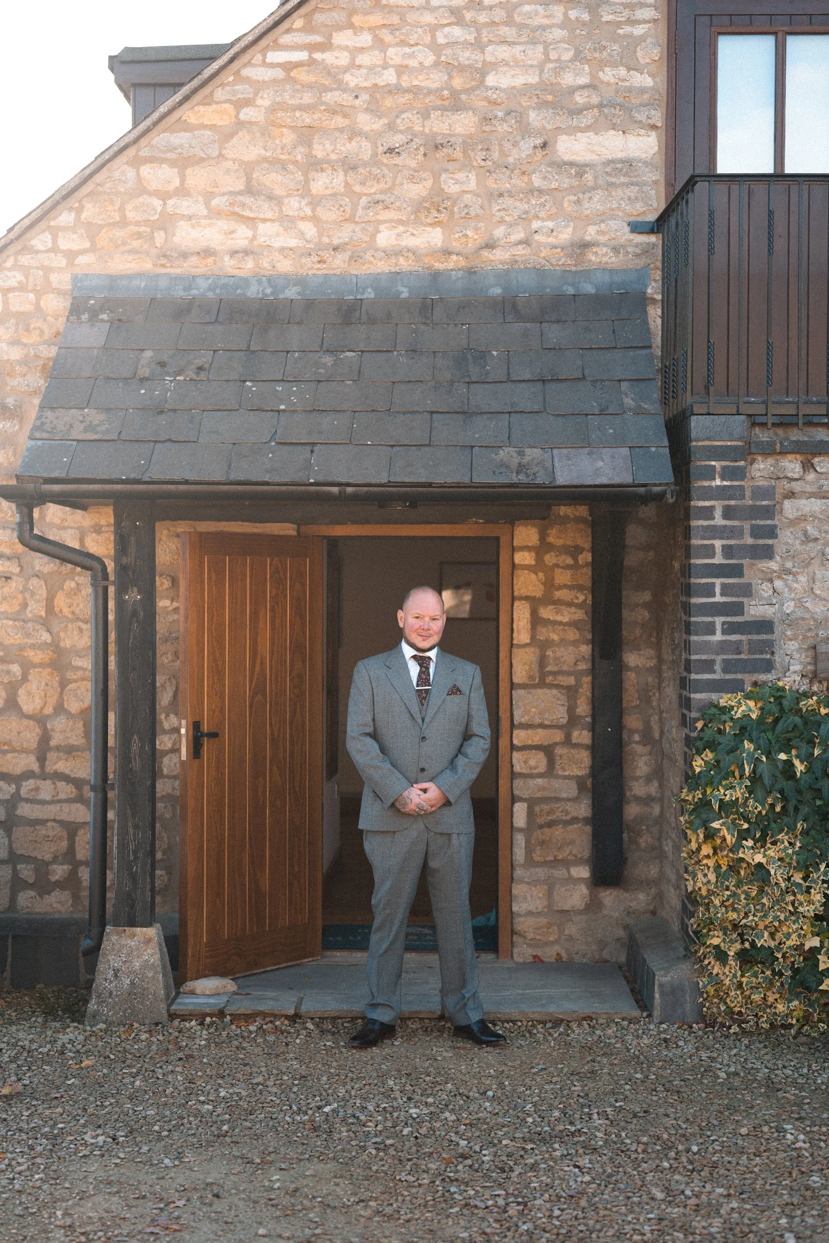 A man in a gray suit with a tie and pocket square, standing at the entrance of a stone house with a wooden door, gravel ground, bushes, and a second story balcony.