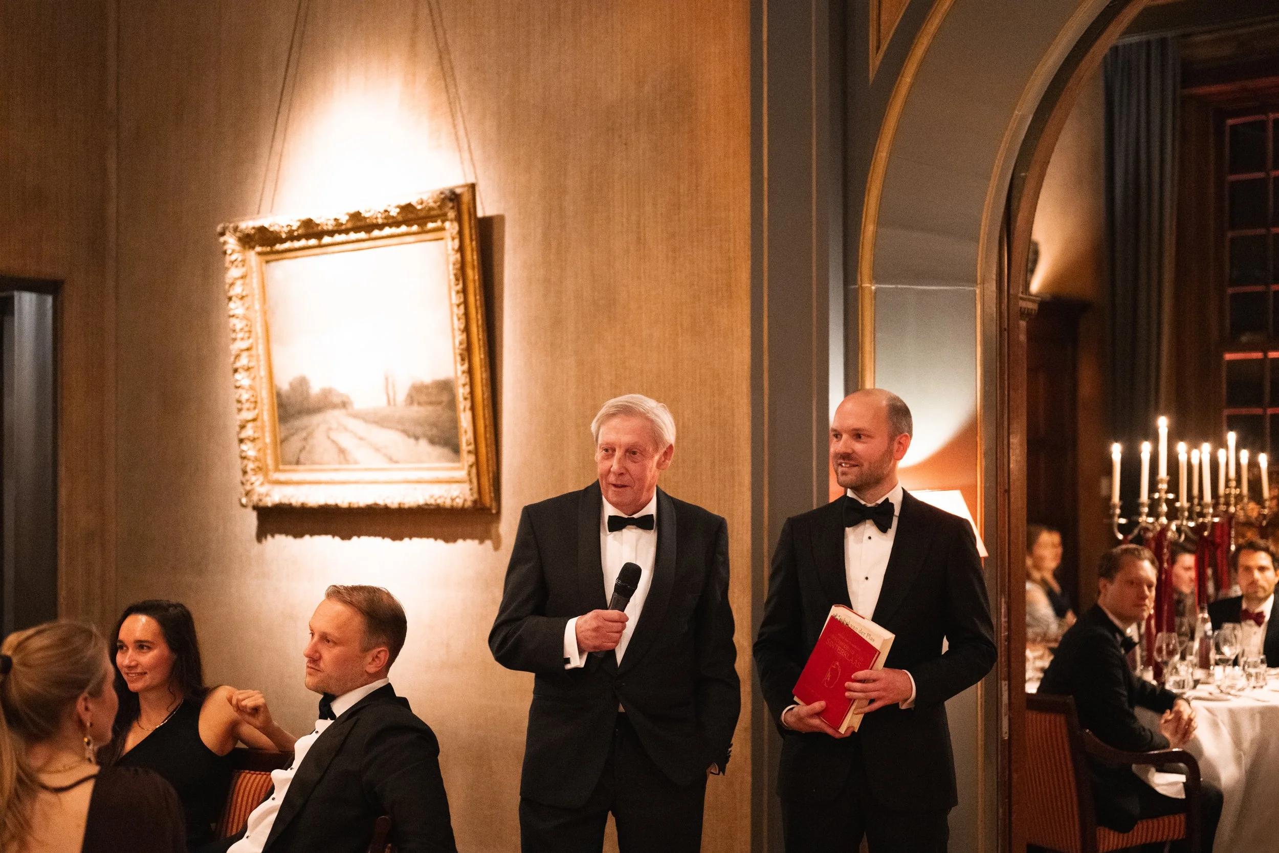 Two men in tuxedos standing and talking during a formal dinner, with multiple guests seated at tables with white tablecloths and candelabras, in an elegant room with warm lighting, a framed landscape painting, and a large mirror reflecting part of th