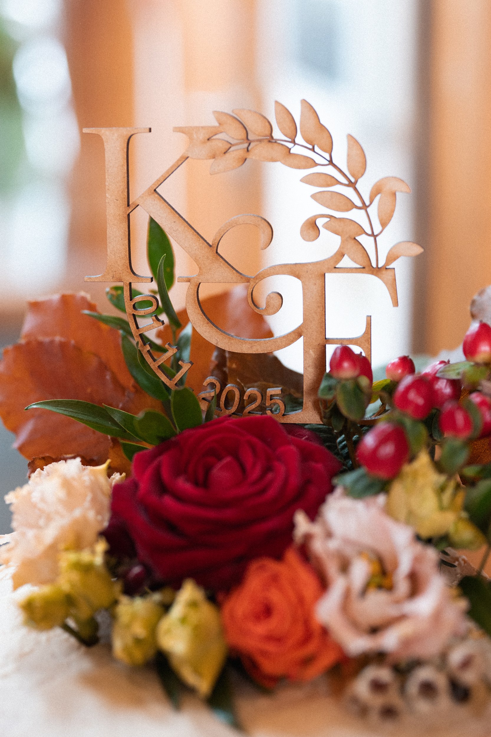 A floral arrangement with red, white, and orange roses, greenery, and berries, topped with a wooden monogram cake topper reading "K & E" with the date 2025.